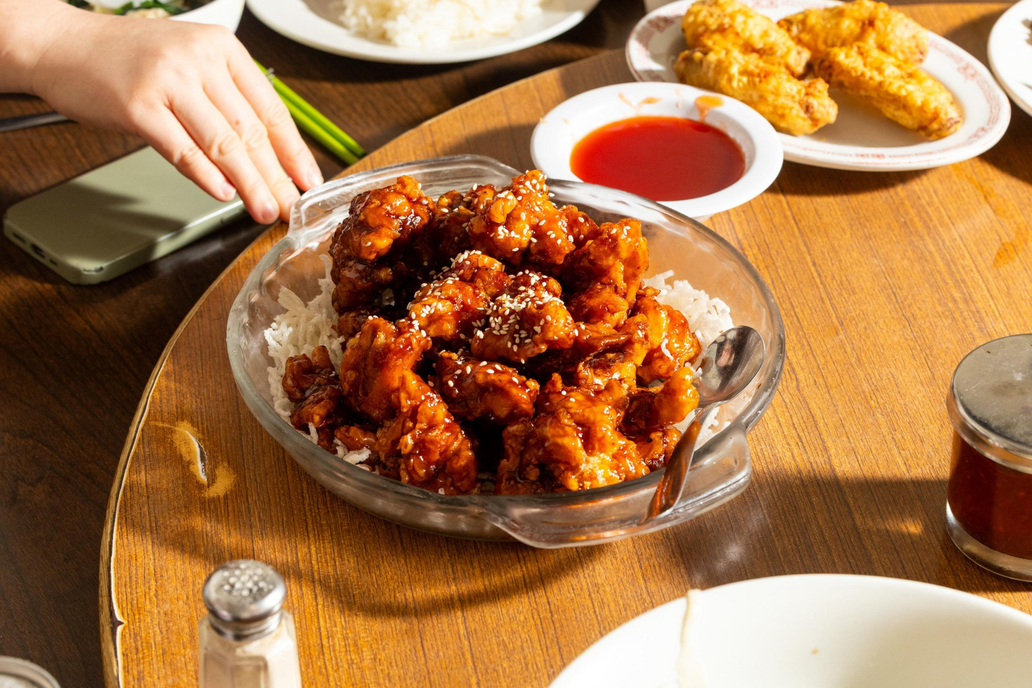 A glass bowl filled with sesame-coated, spicy fried chicken on white rice sits on a wooden table surrounded by plates of fried chicken, a bowl of red sauce, and a hand reaching for chopsticks.