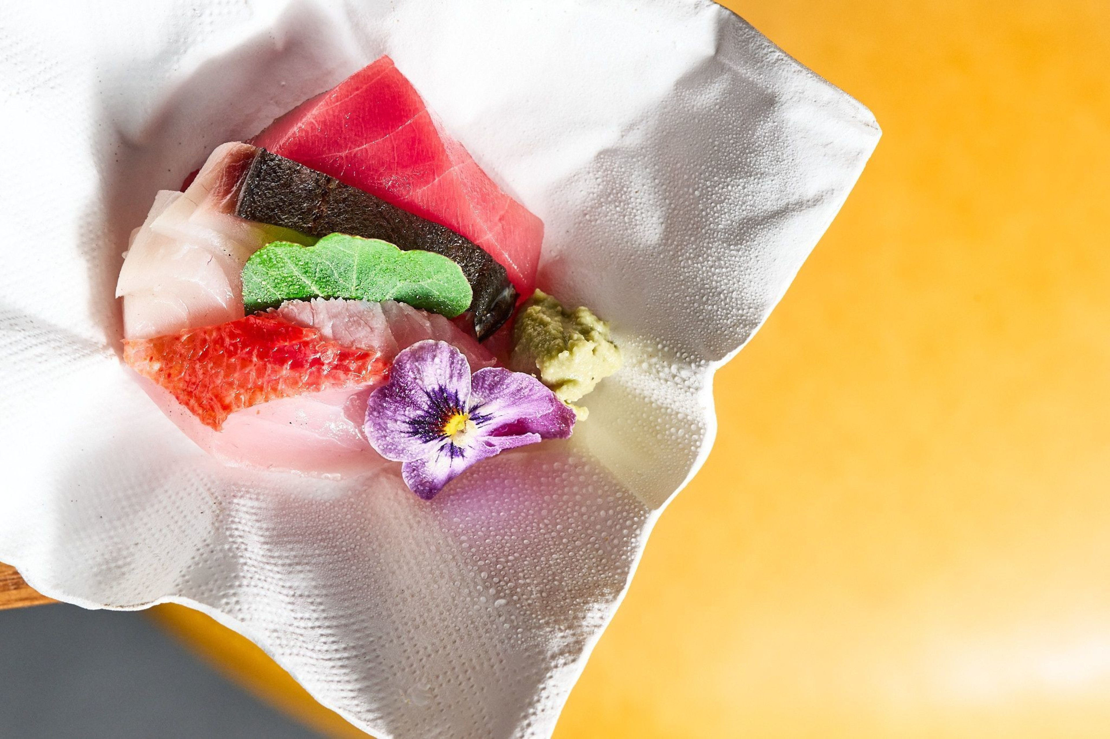The image shows assorted sashimi elegantly arranged with a purple flower and wasabi, all placed on a white textured cloth against a golden yellow background.