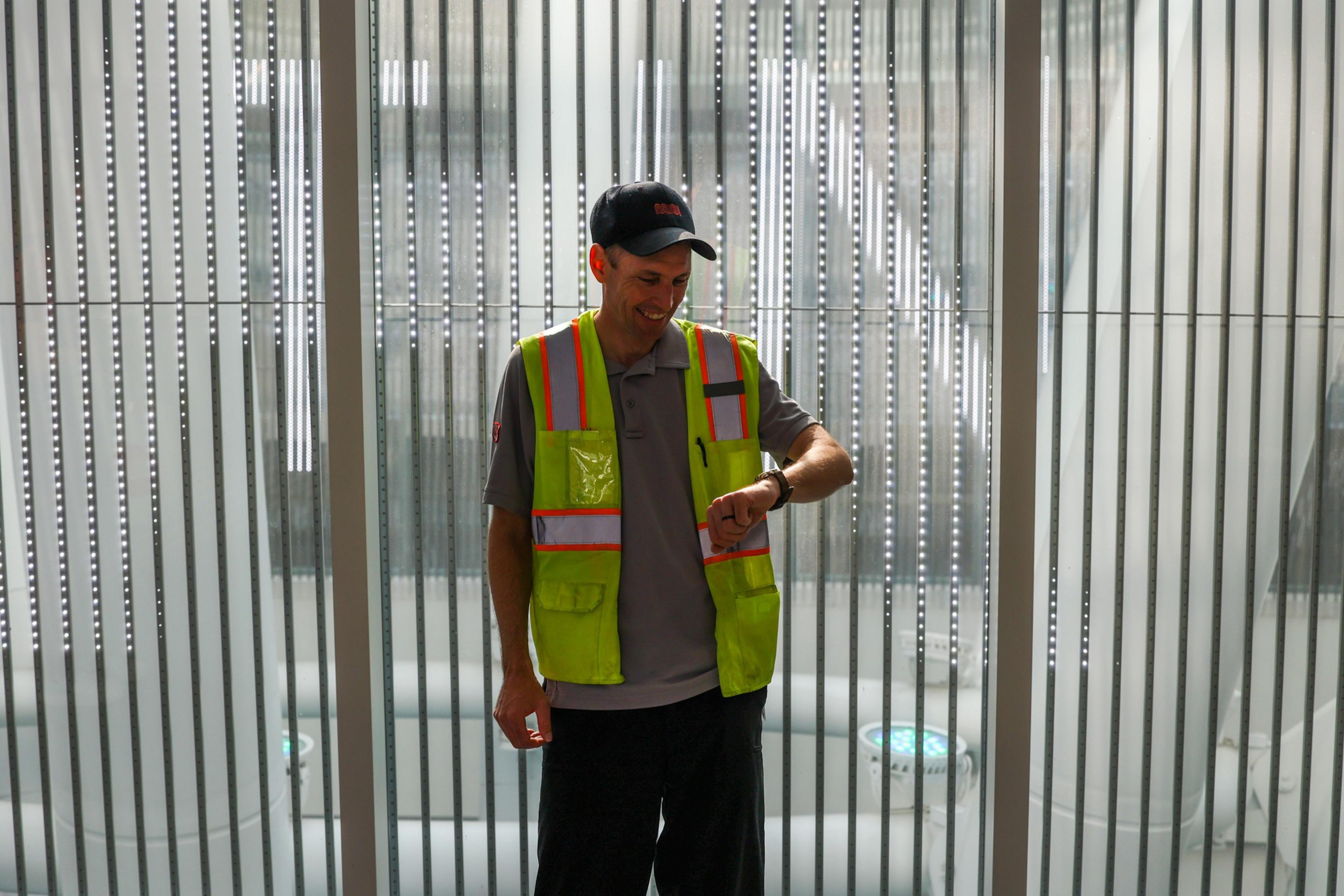 A person wearing a neon safety vest and cap is looking at their wristwatch. They are standing in front of a striped, metallic background.