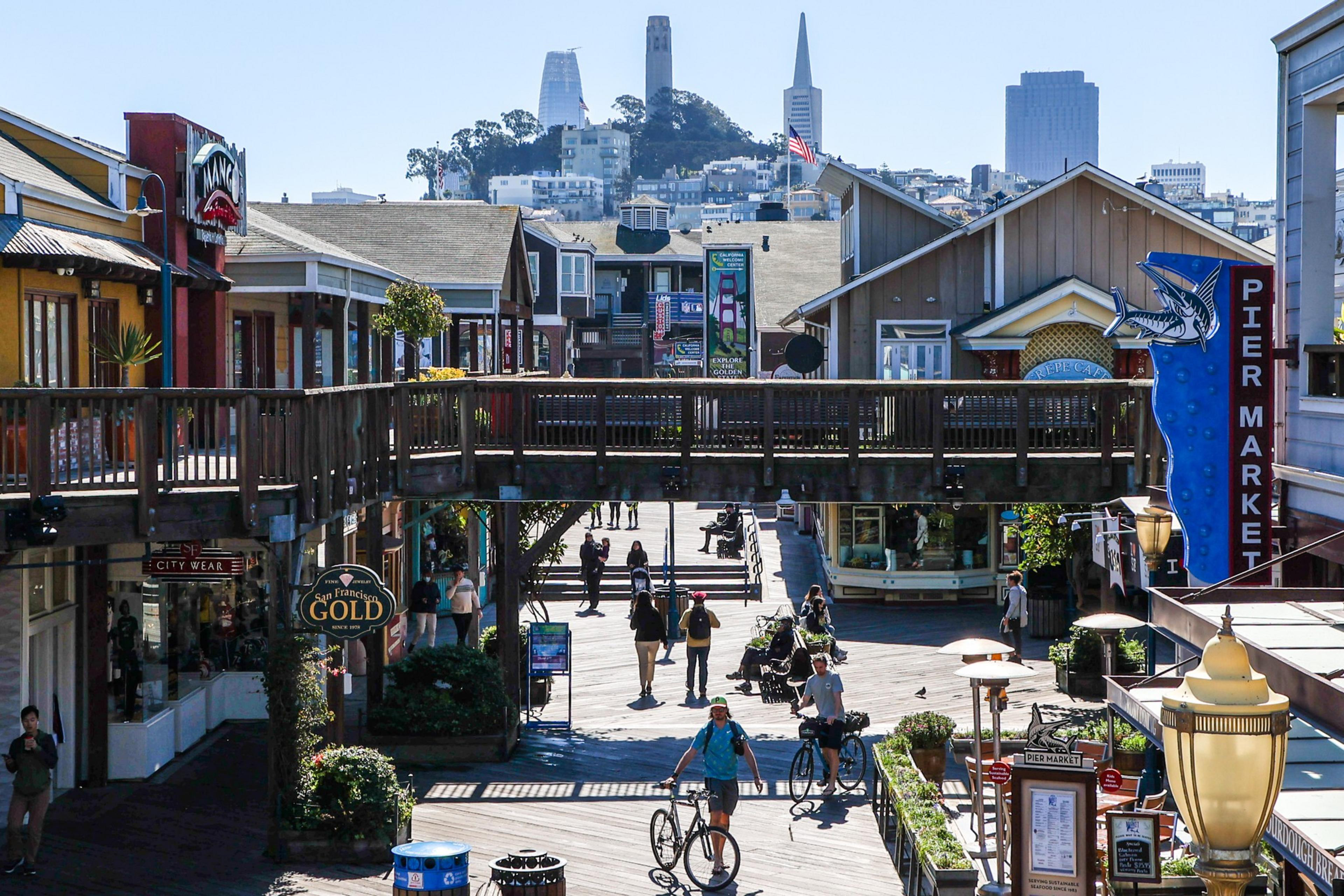 A bustling pier market with wooden walkways, shops, people walking and biking, and a city skyline with Coit Tower and Transamerica Pyramid in the background.
