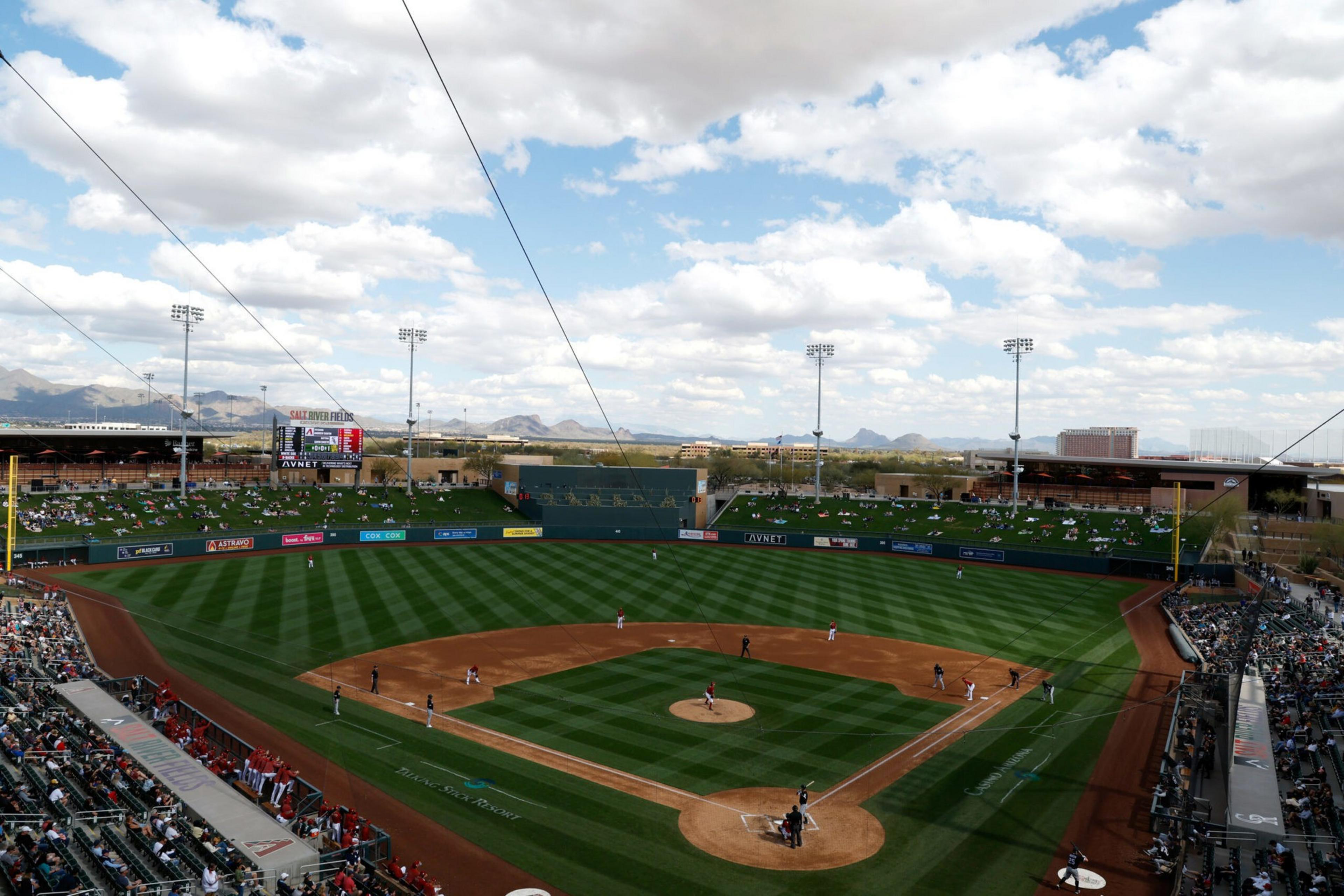 A baseball field with players in position, spectators in the stands, a scoreboard, bright daylight, and mountains visible under a cloudy sky.