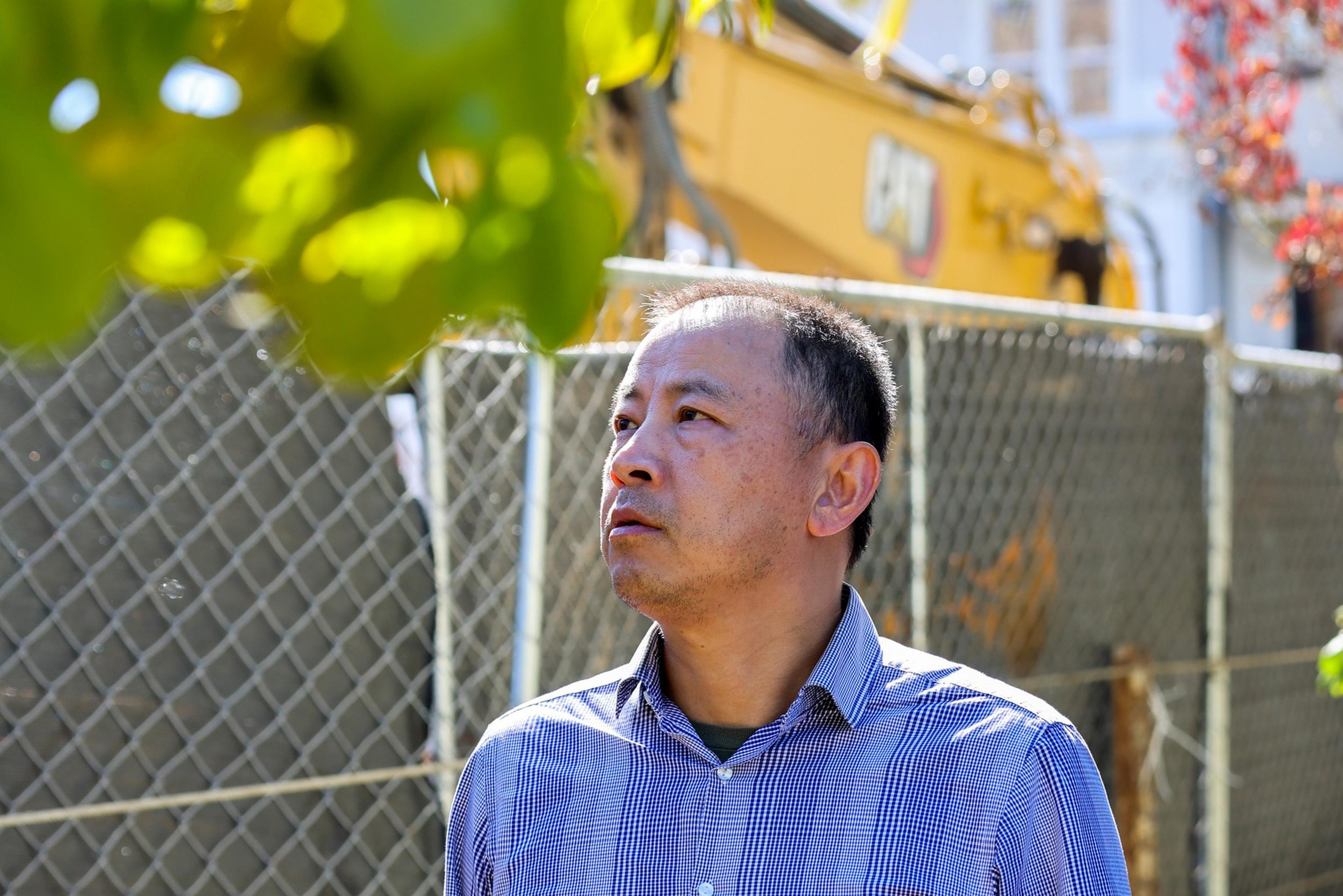 A man in a checkered shirt looks thoughtful, standing outdoors by a chain-link fence, with construction equipment and blurred foliage in the background.