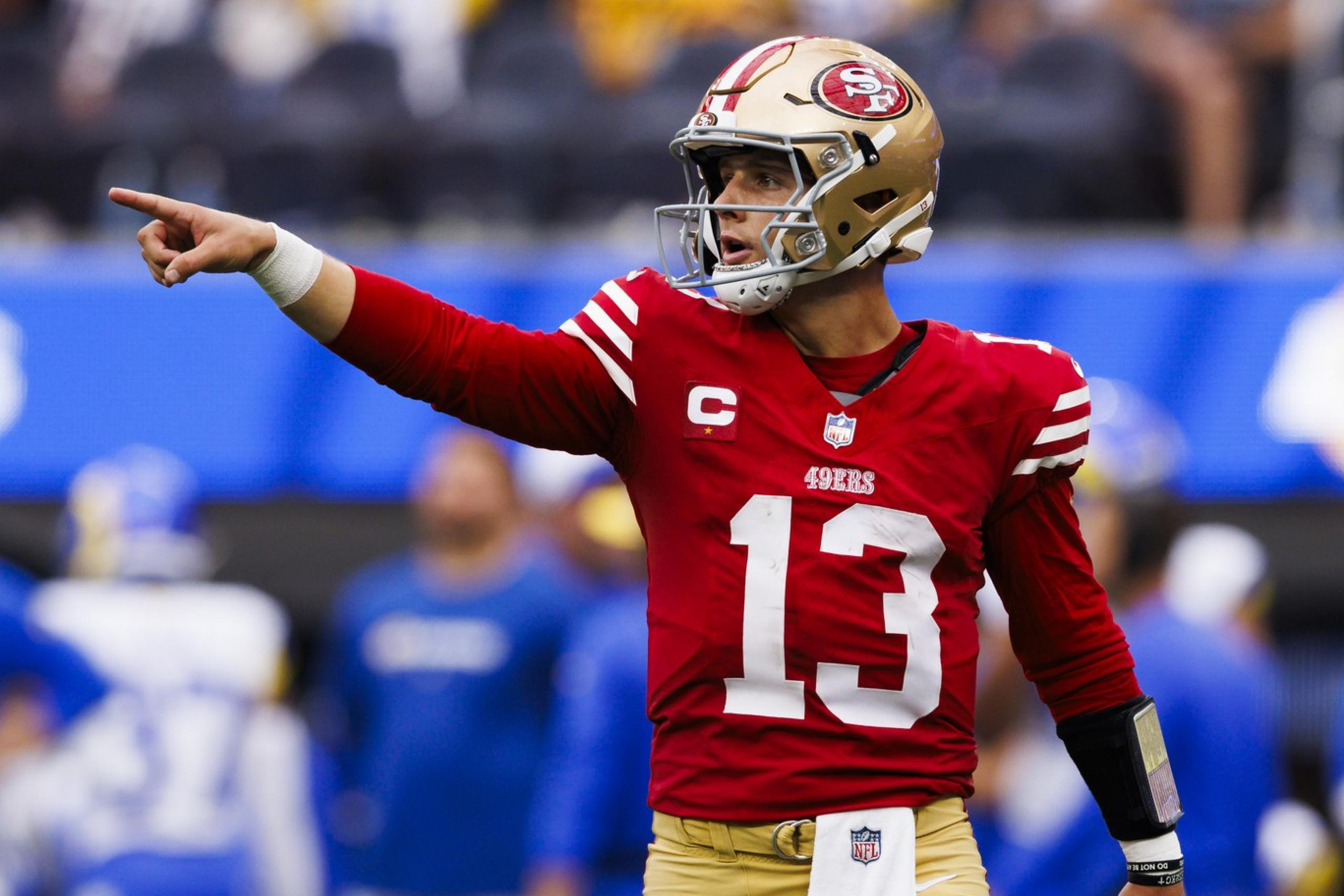 A football player in a red jersey and gold pants is on the field, pointing with his right arm extended. He wears a helmet and wristbands with a white towel tucked in.