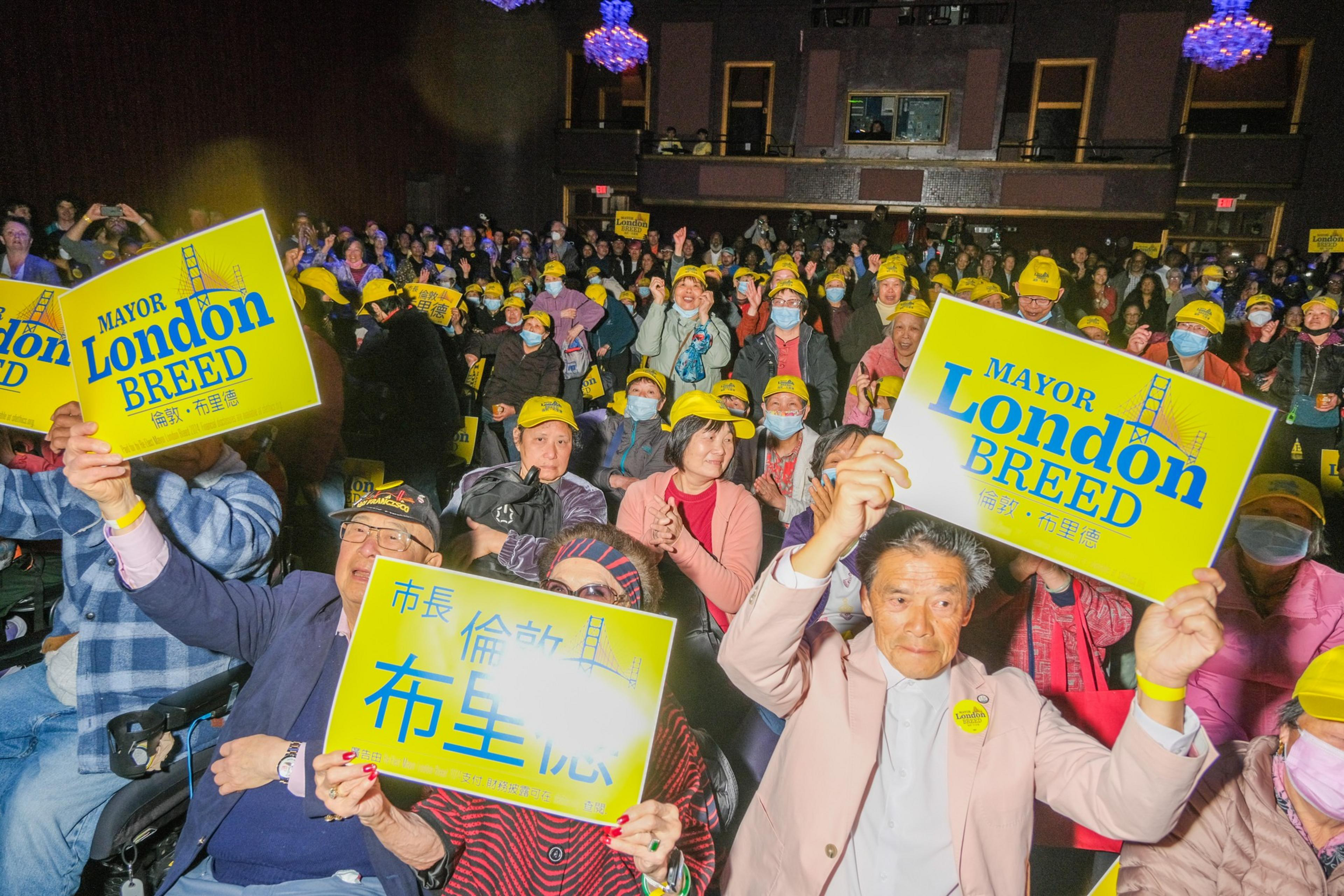 A crowd of joyful people, many wearing yellow hats, holds up signs supporting Mayor London Breed, in a well-lit indoor setting.