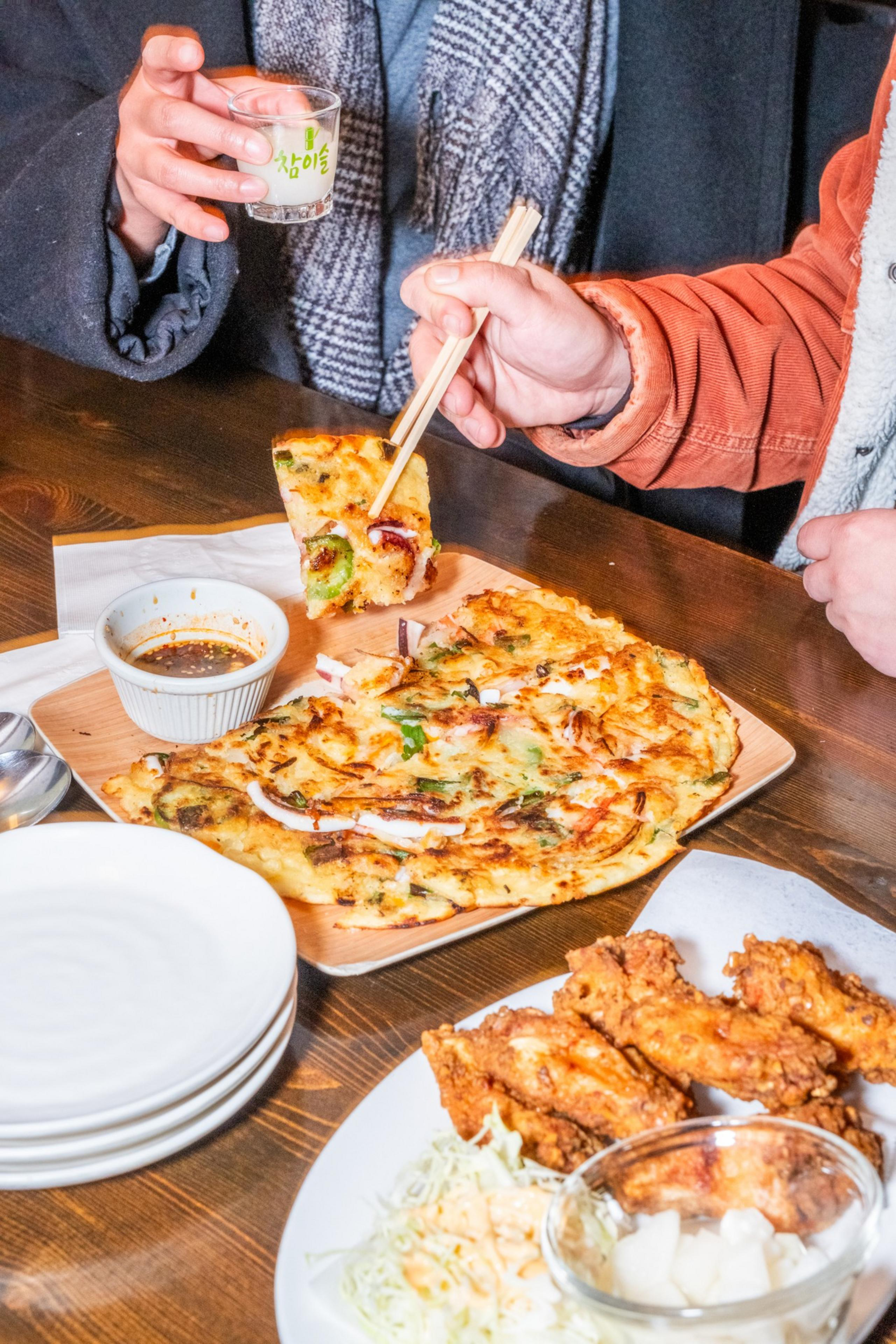 Two people are enjoying a meal. One holds a glass, the other uses chopsticks to pick up a piece of savory pancake with dipping sauce nearby.