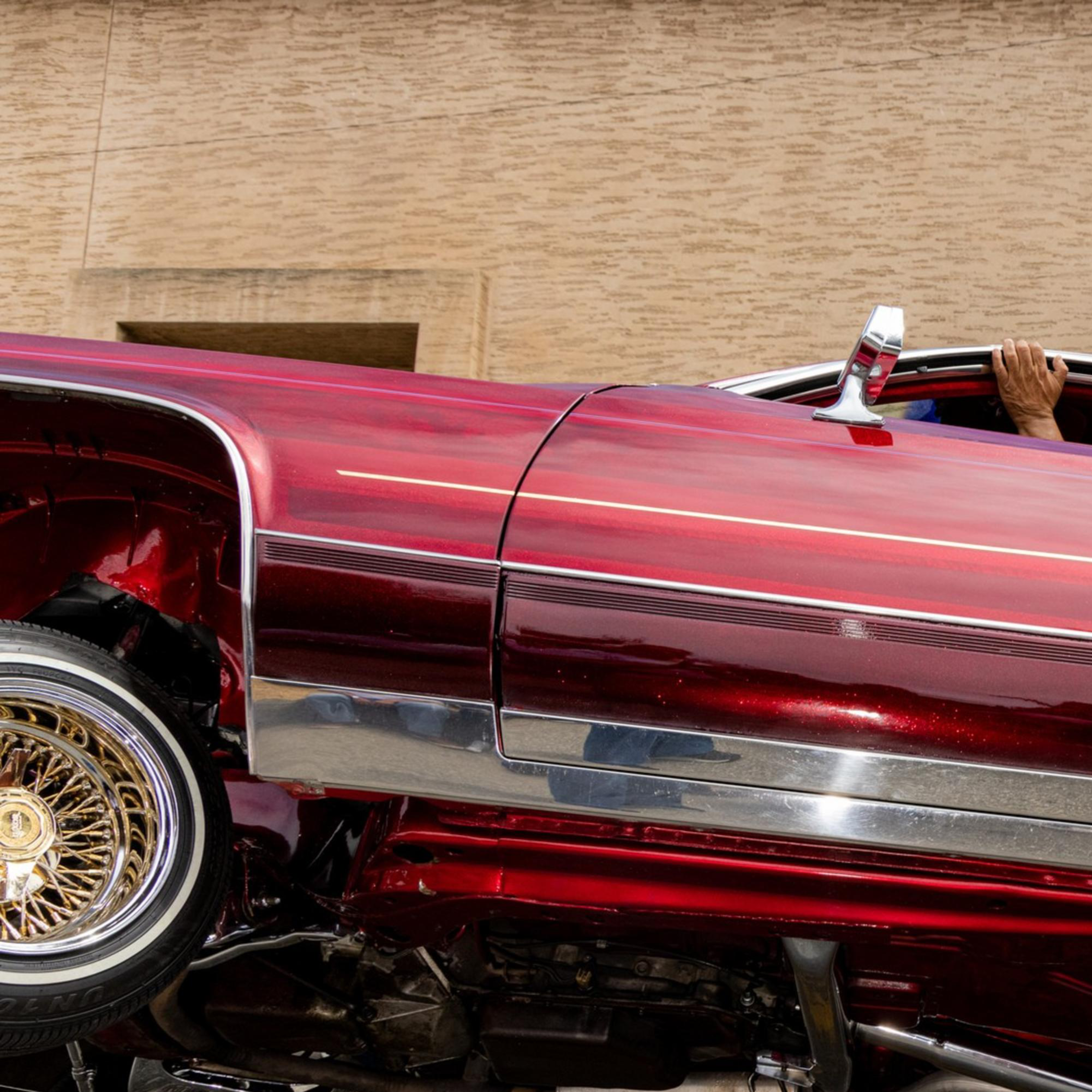 A red lowrider car with shiny chrome details is tilted diagonally, showing its undercarriage. A hand is visible from the rolled-down window, gripping it.