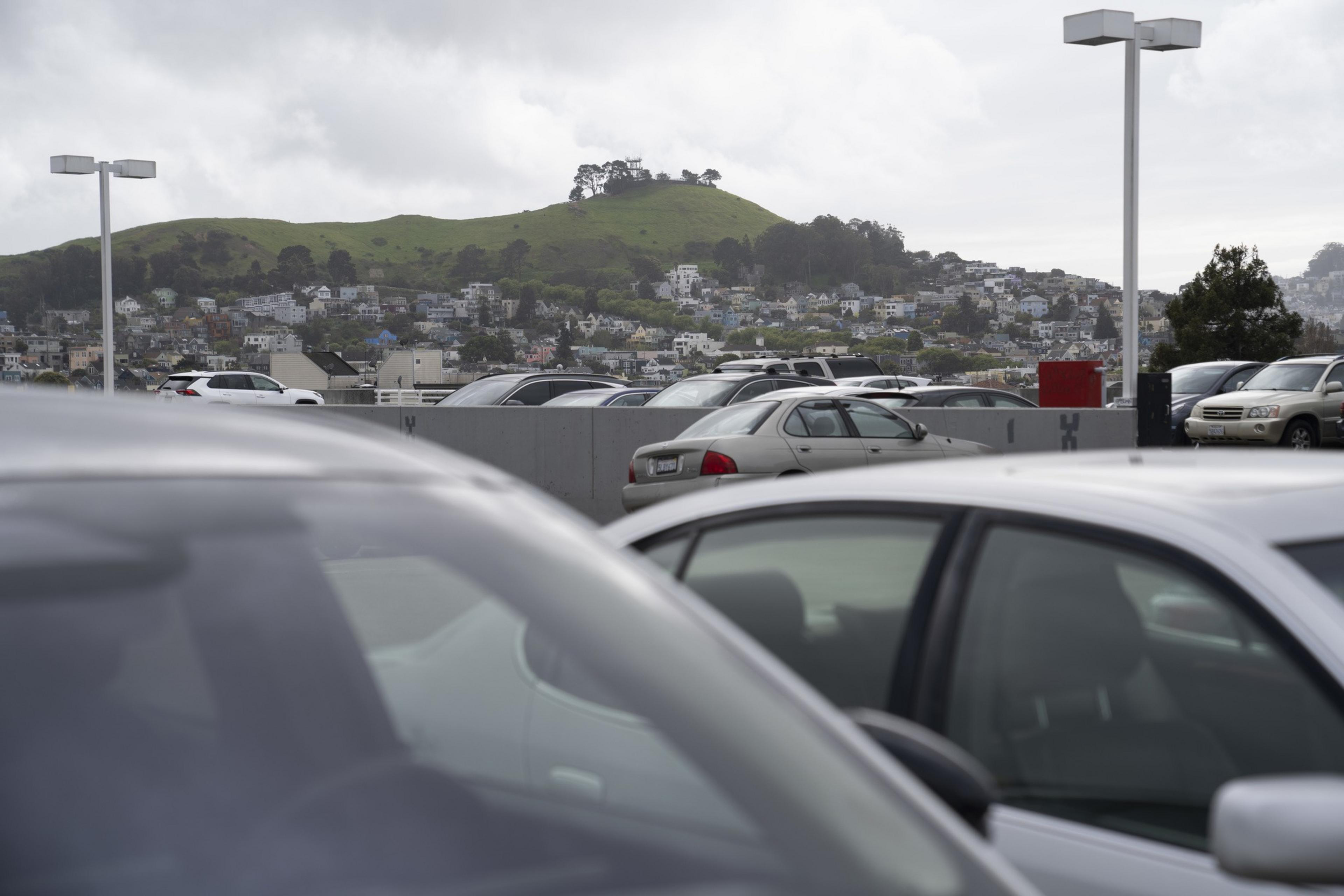 A parking lot filled with cars is in the foreground, while a grassy hill and a residential area with houses are visible in the background under a cloudy sky.