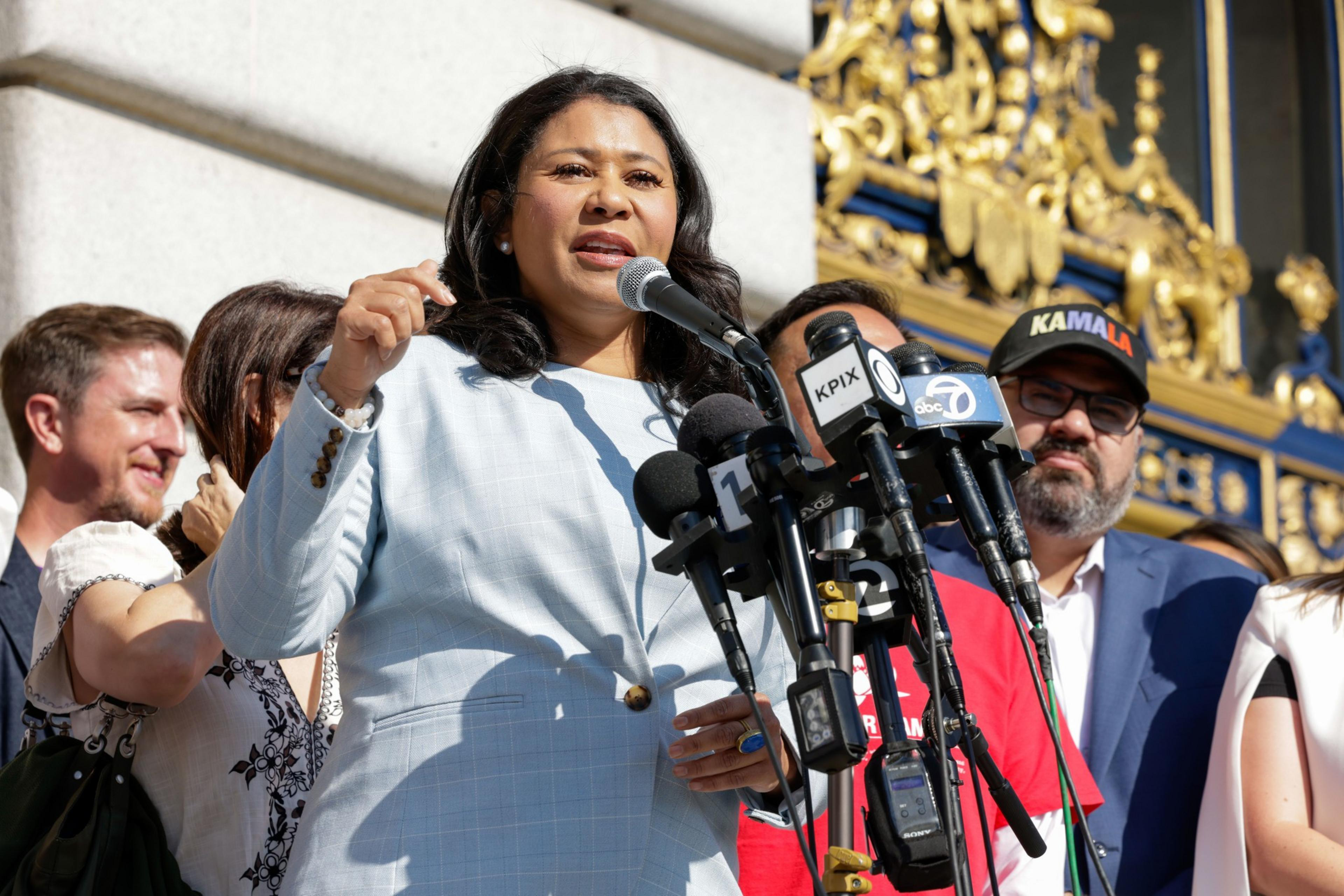 A woman in a light blue suit is speaking at a podium with multiple microphones. She is surrounded by several people, and intricate gold decor can be seen in the background.