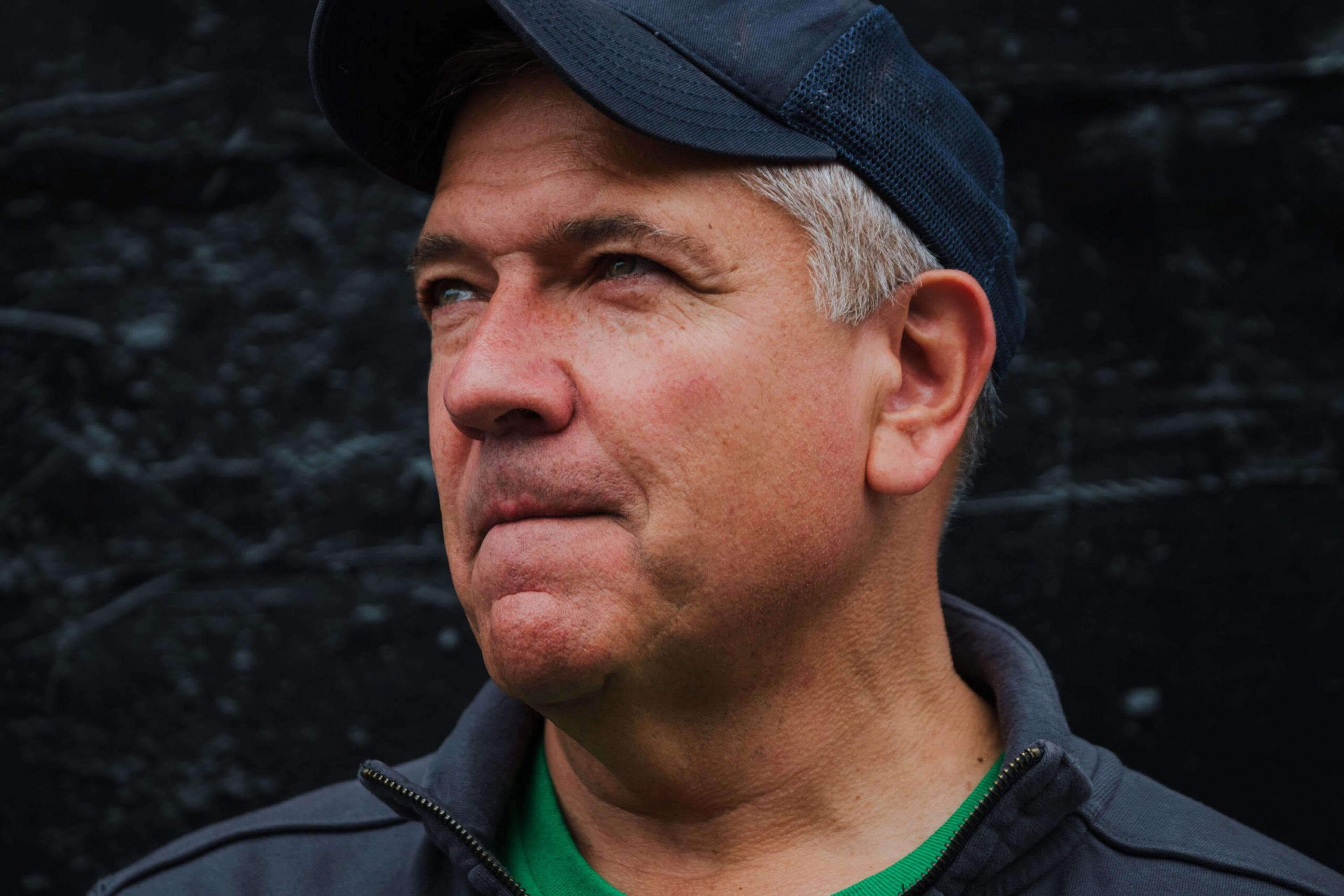 A middle-aged man with gray hair wears a dark cap and jacket, looking thoughtfully to the side against a dark, textured background.