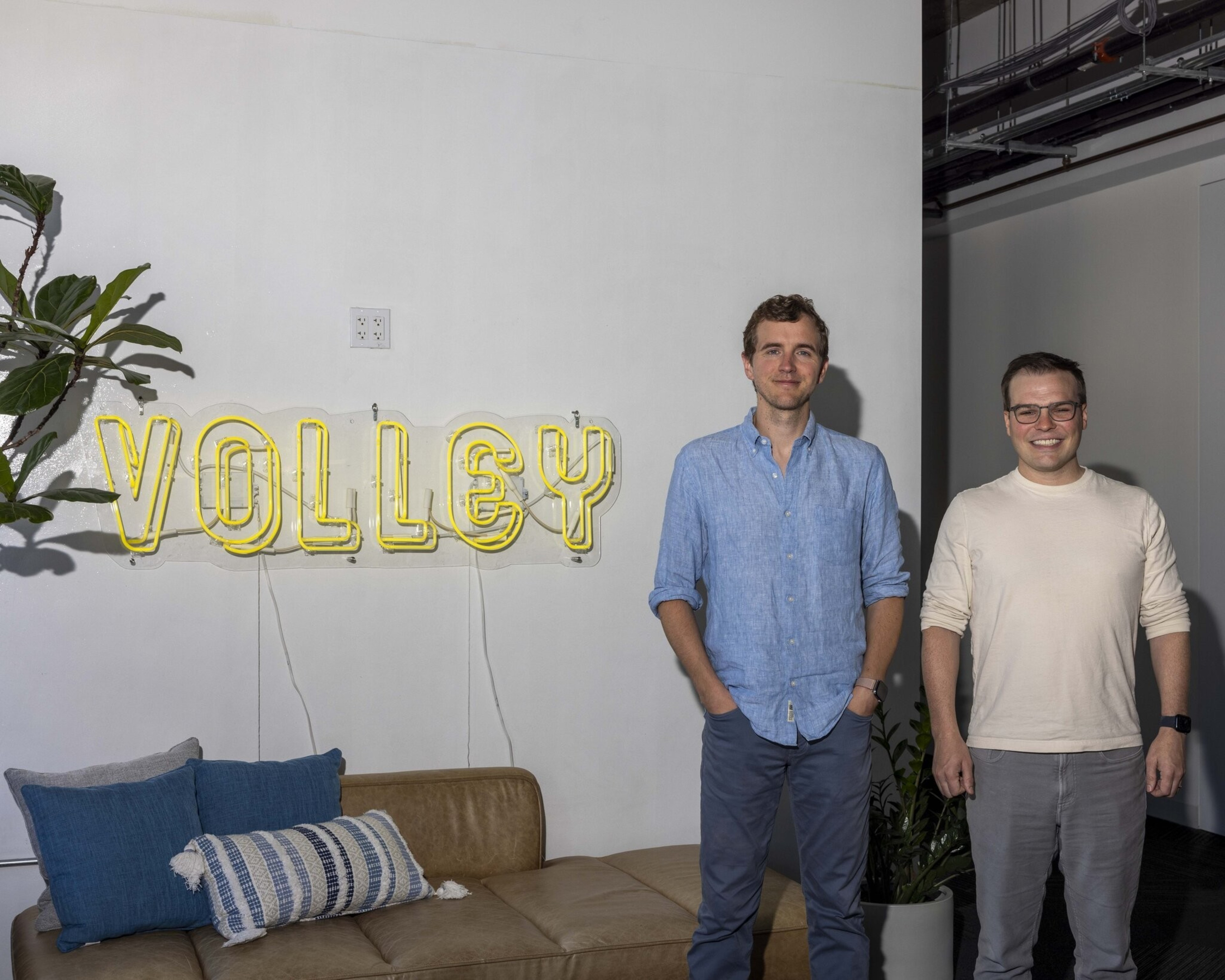 Two men stand in front of a wall, above a tan couch and beside a plant. The wall has a neon sign that reads "VOLLEY" in yellow letters.