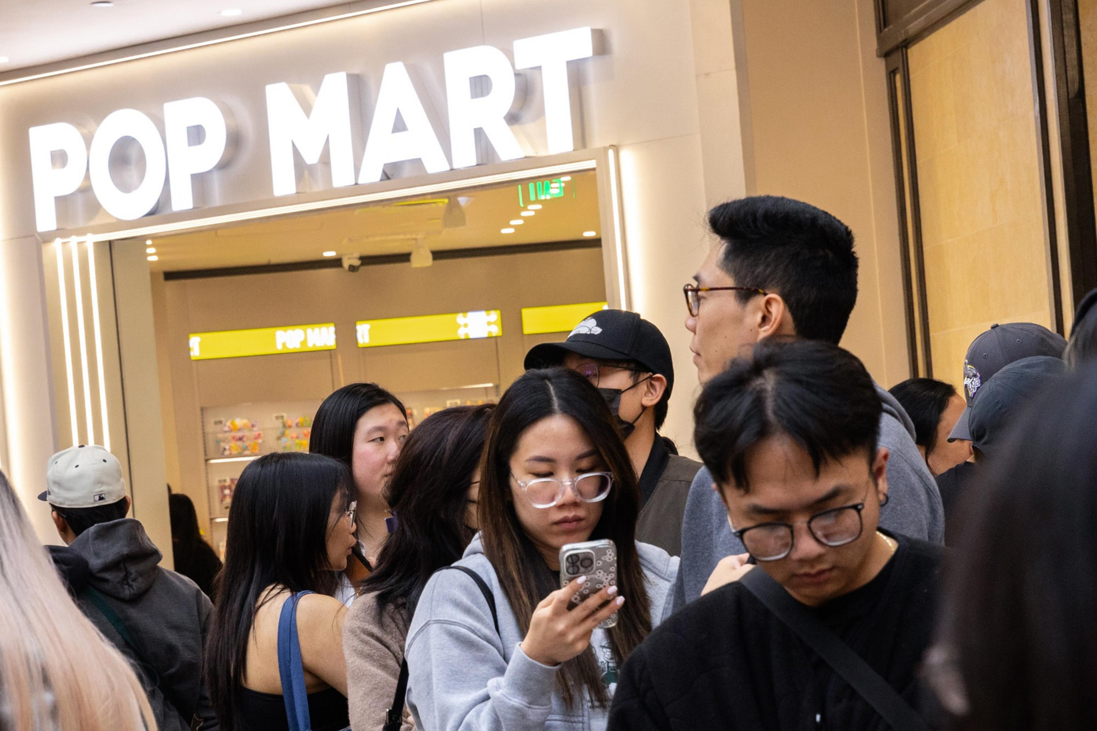 People are gathered in front of a store named "POP MART", with many looking at their phones while standing in line.