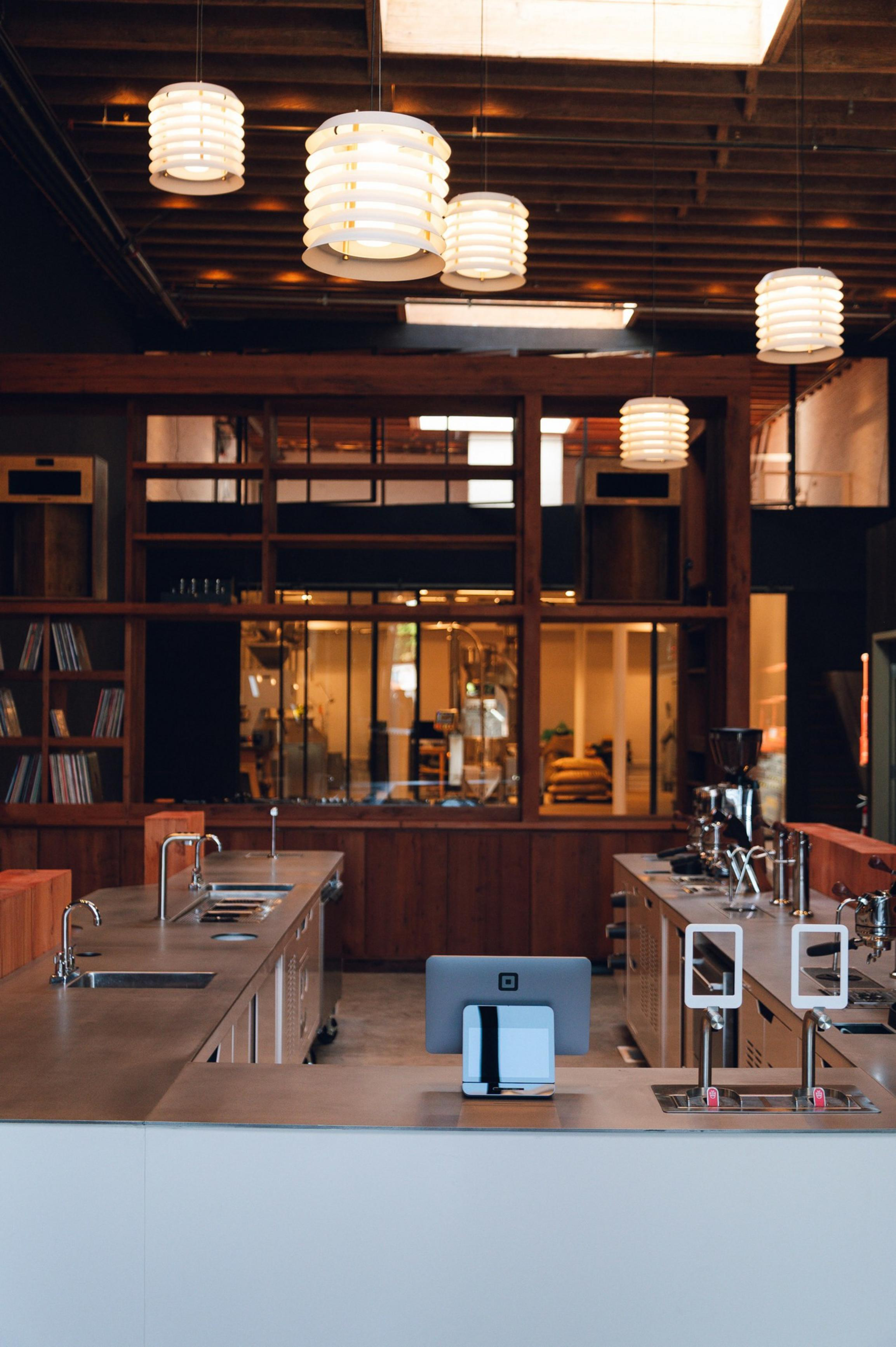 A modern kitchen with stainless steel counters, sinks, coffee equipment, hanging pendant lights, and wooden shelves in the background.