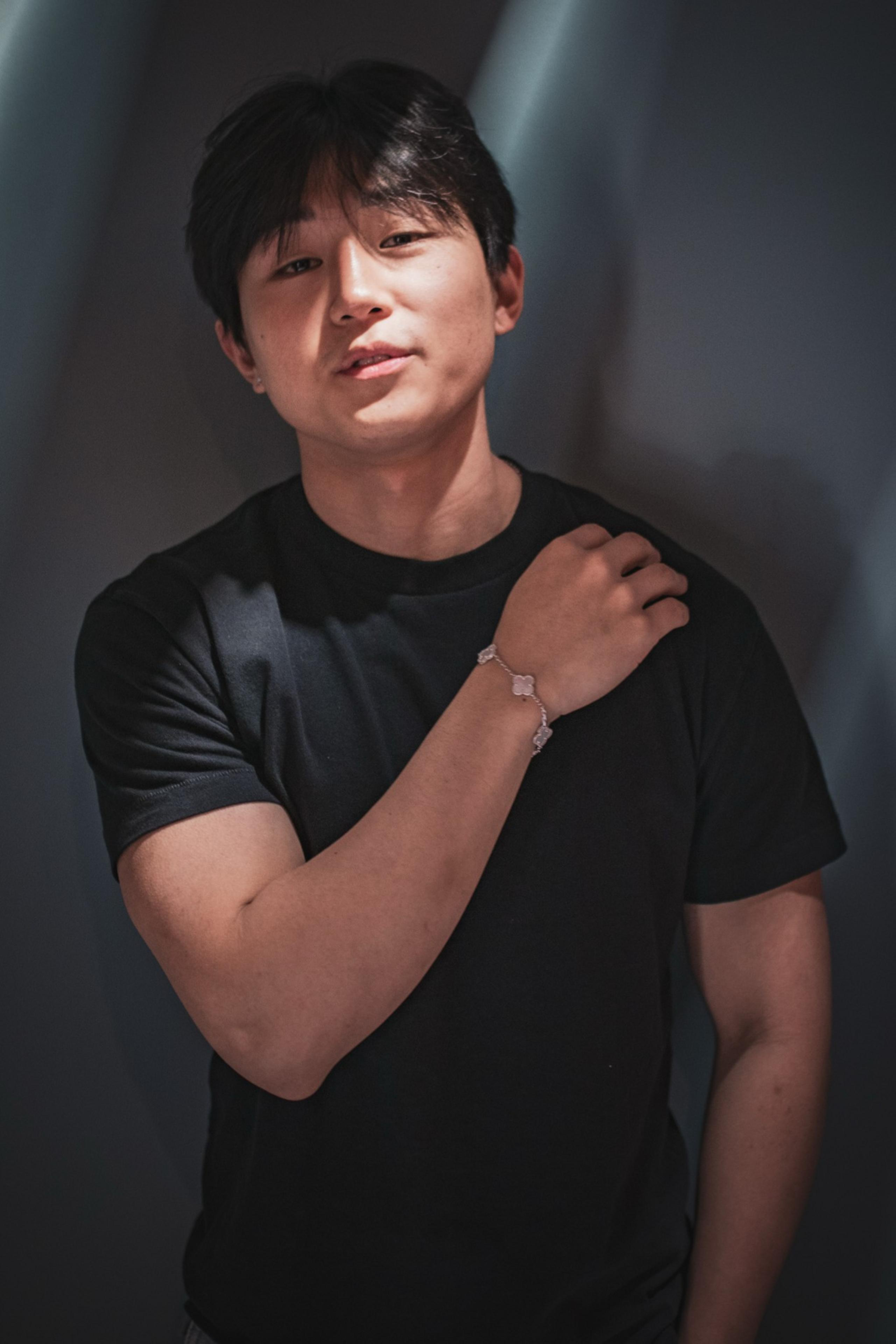 A person with dark hair is wearing a black t-shirt and a bracelet, resting one hand on their shoulder. The lighting is soft and shadowy.