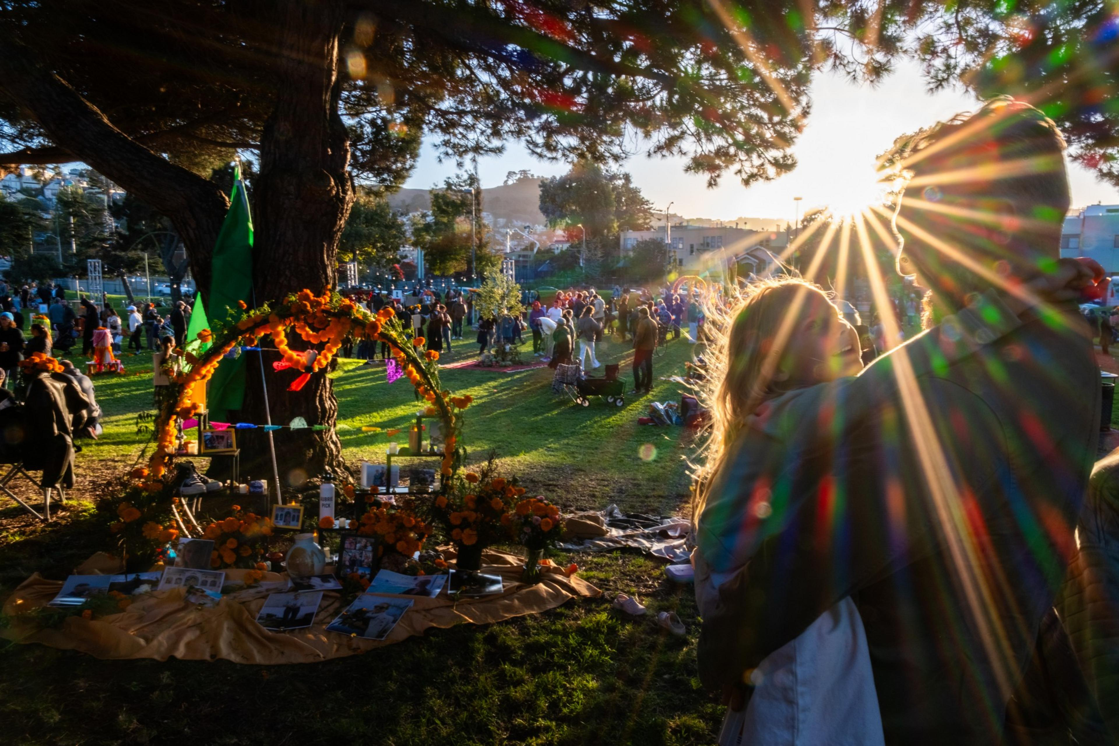 A man and child embrace in a sunlit park with a decorated altar of flowers and photographs. People gather around, creating a festive and warm atmosphere.