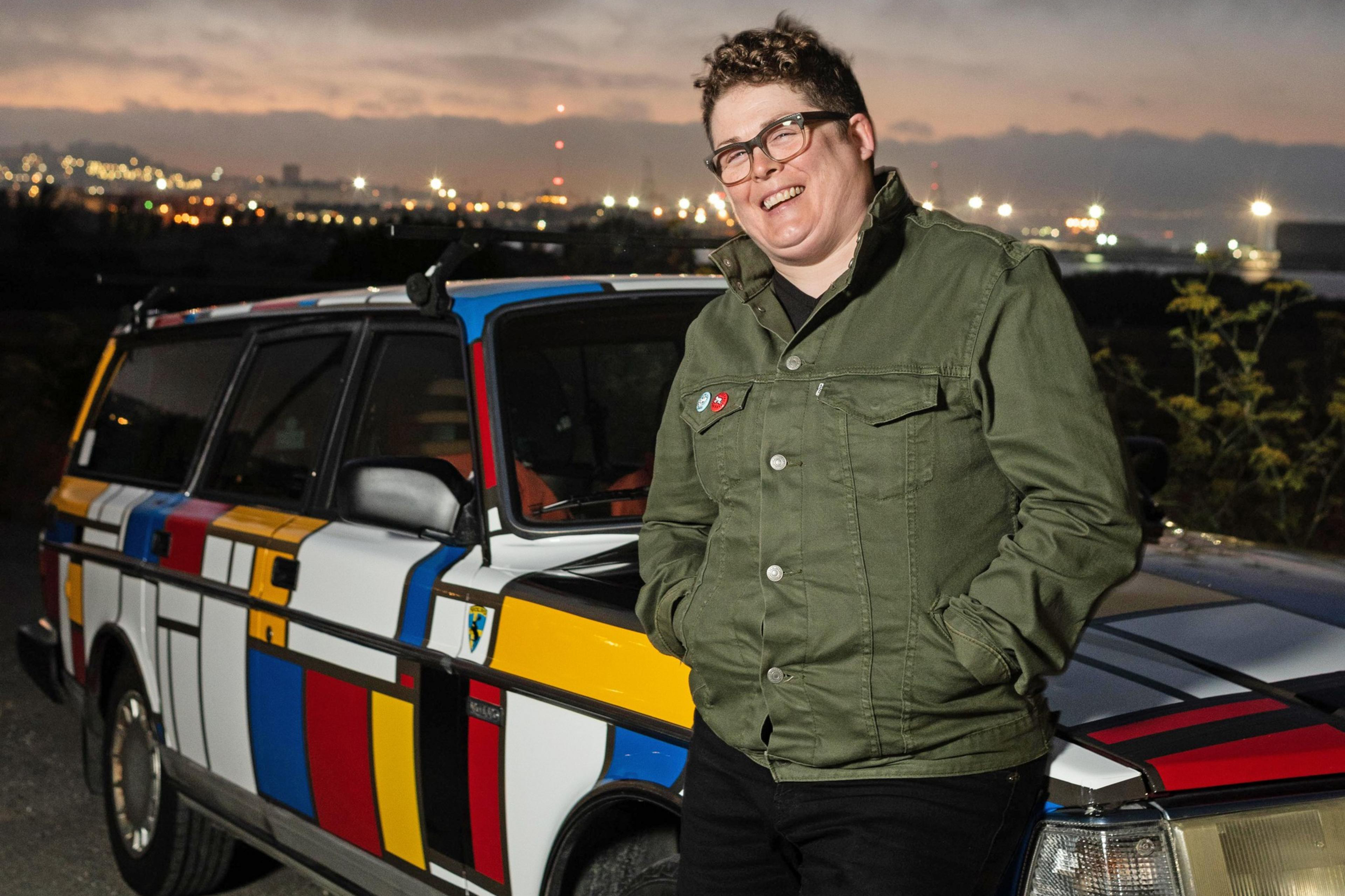 A person wearing glasses and a green jacket leans against a vibrantly patterned car with a geometric design of red, blue, yellow, and black lines at dusk.