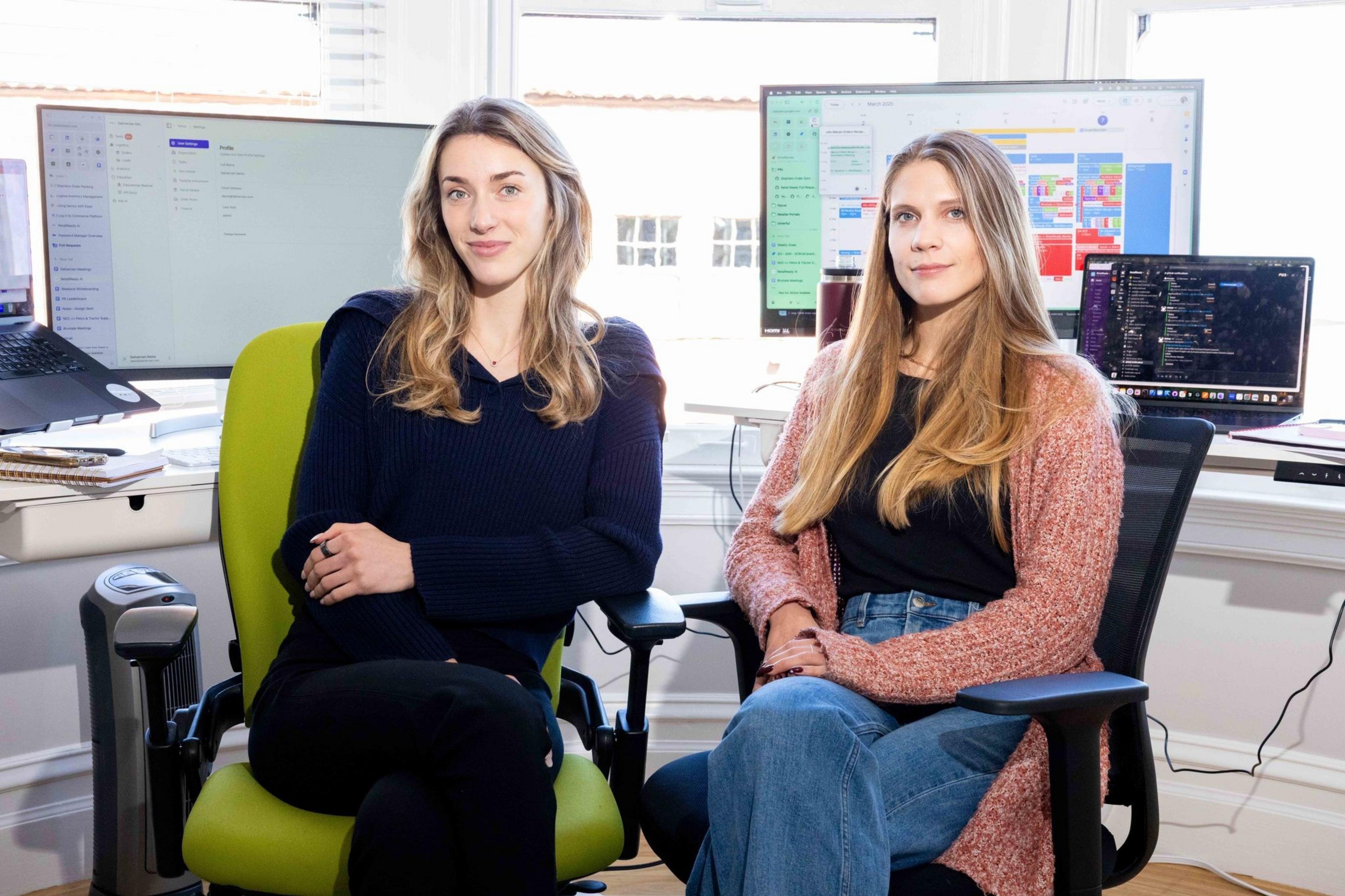 Two women are seated in an office environment. They are sitting in chairs with computer screens and laptops displaying calendars and messages in the background.