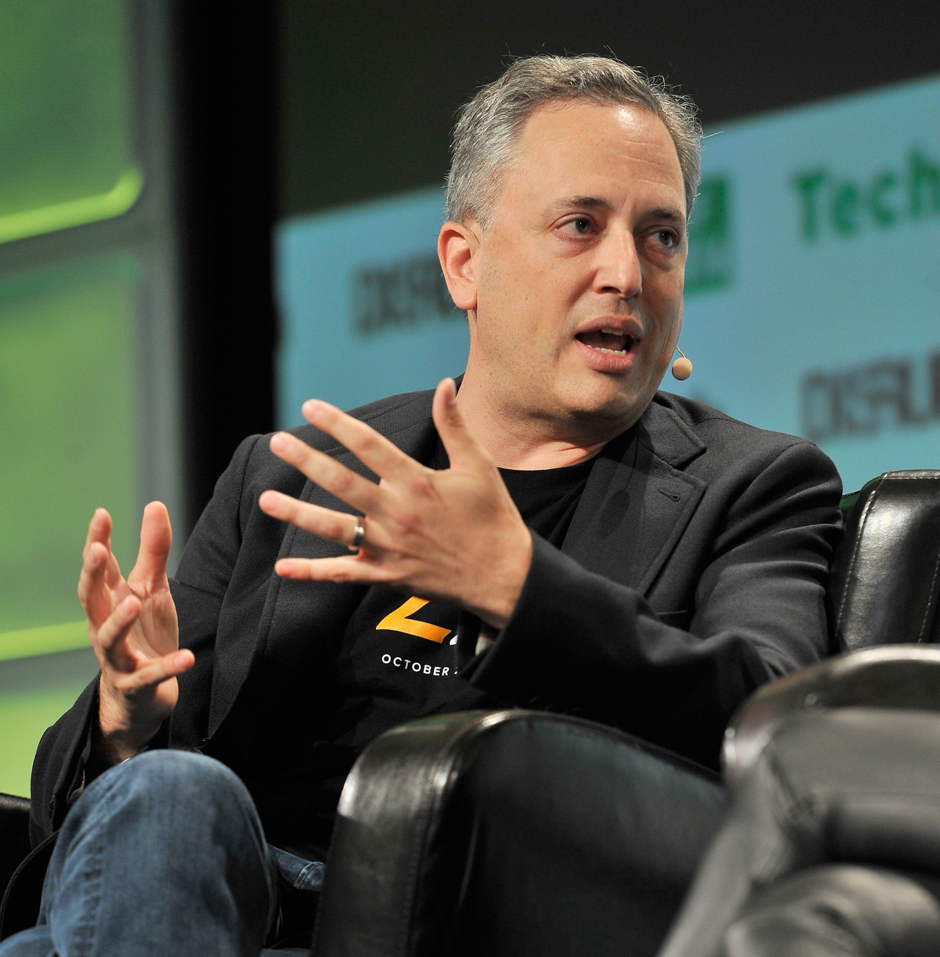 A man with short gray hair is speaking, wearing a black blazer over a black shirt. He is sitting on a black leather armchair, gesturing with his hands animatedly.