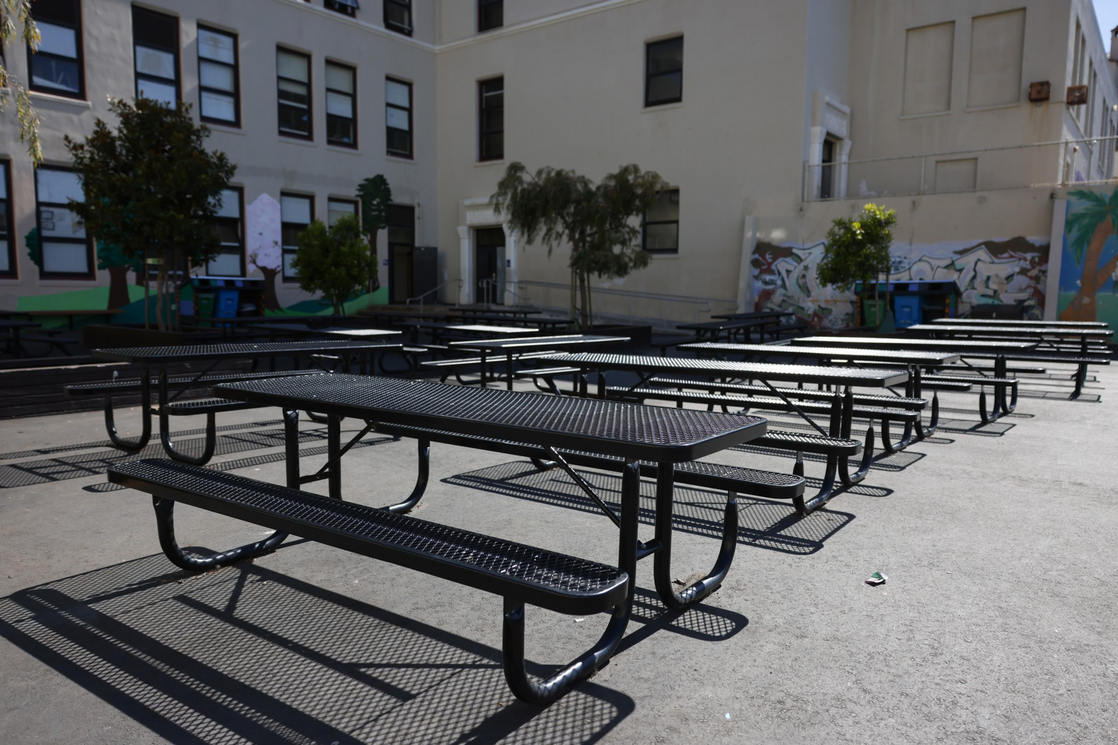 Several empty black metal picnic tables are arranged on a concrete surface outside a cream-colored building with small trees and wall murals nearby.