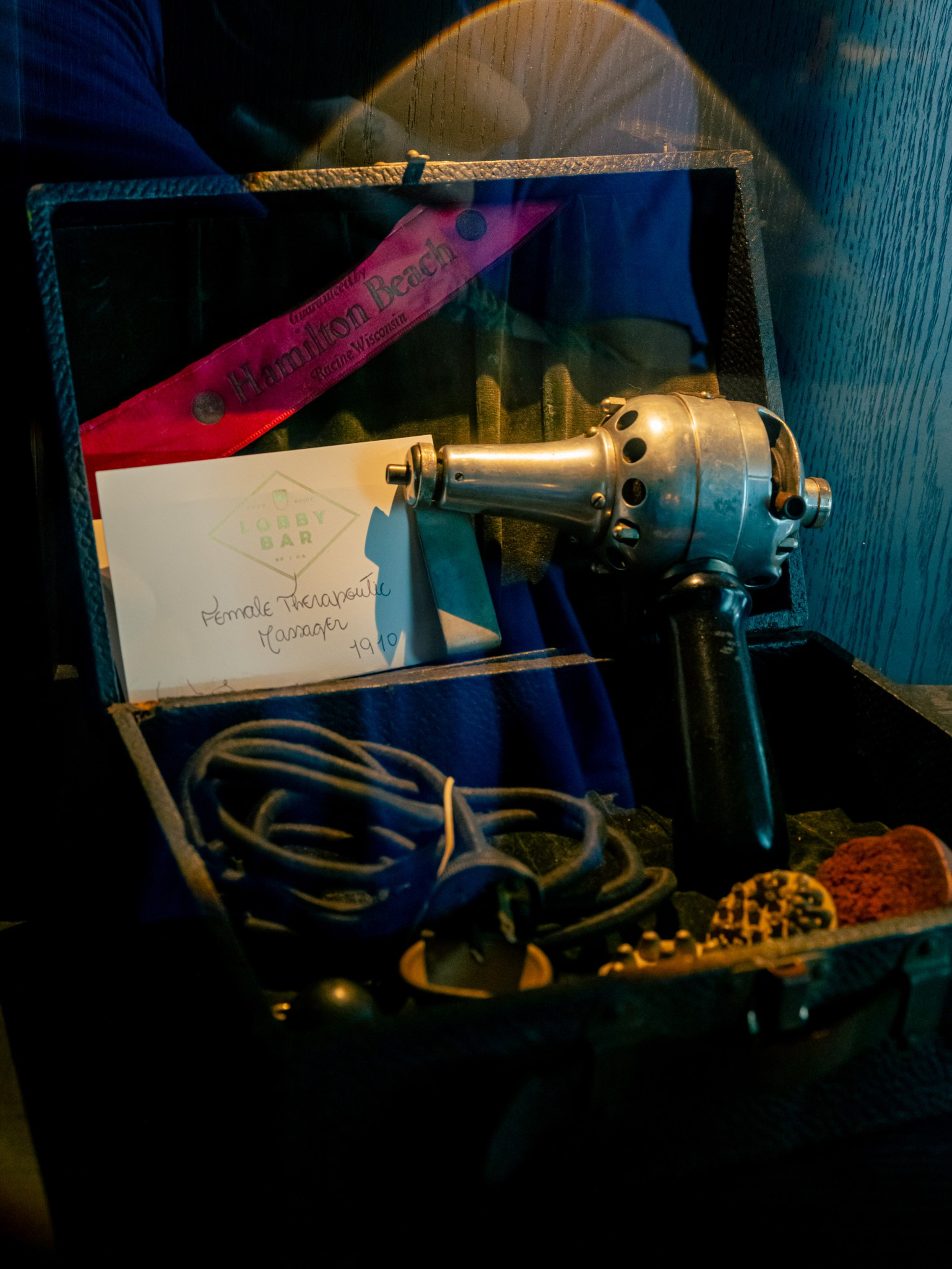 An old metal therapeutic massager with a black handle, cords, and a red sash labeled “Hamilton Beach” is displayed inside an open black case.