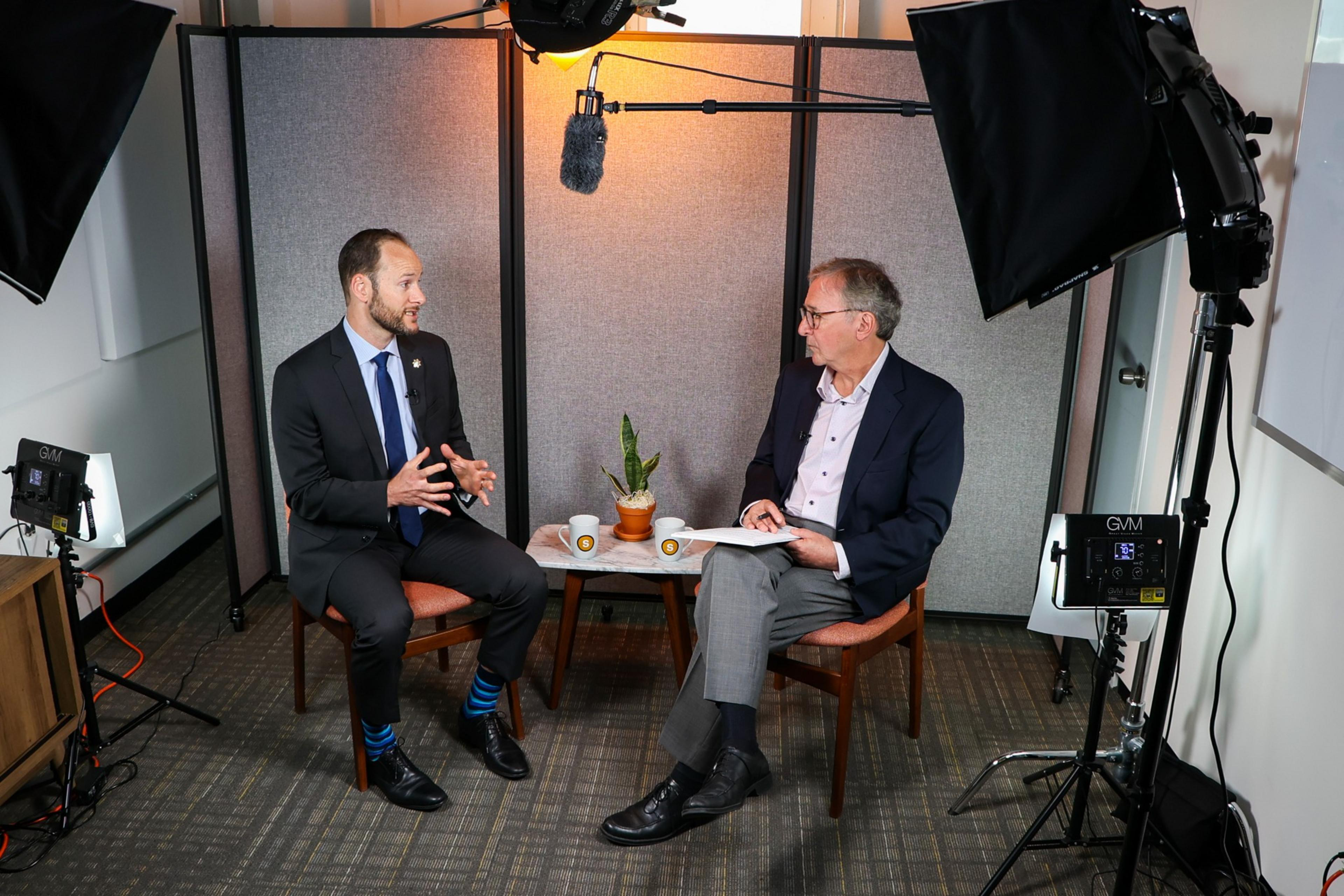 Two men in suits sit opposite each other, having a discussion with microphones and studio lights around them in a small room.
