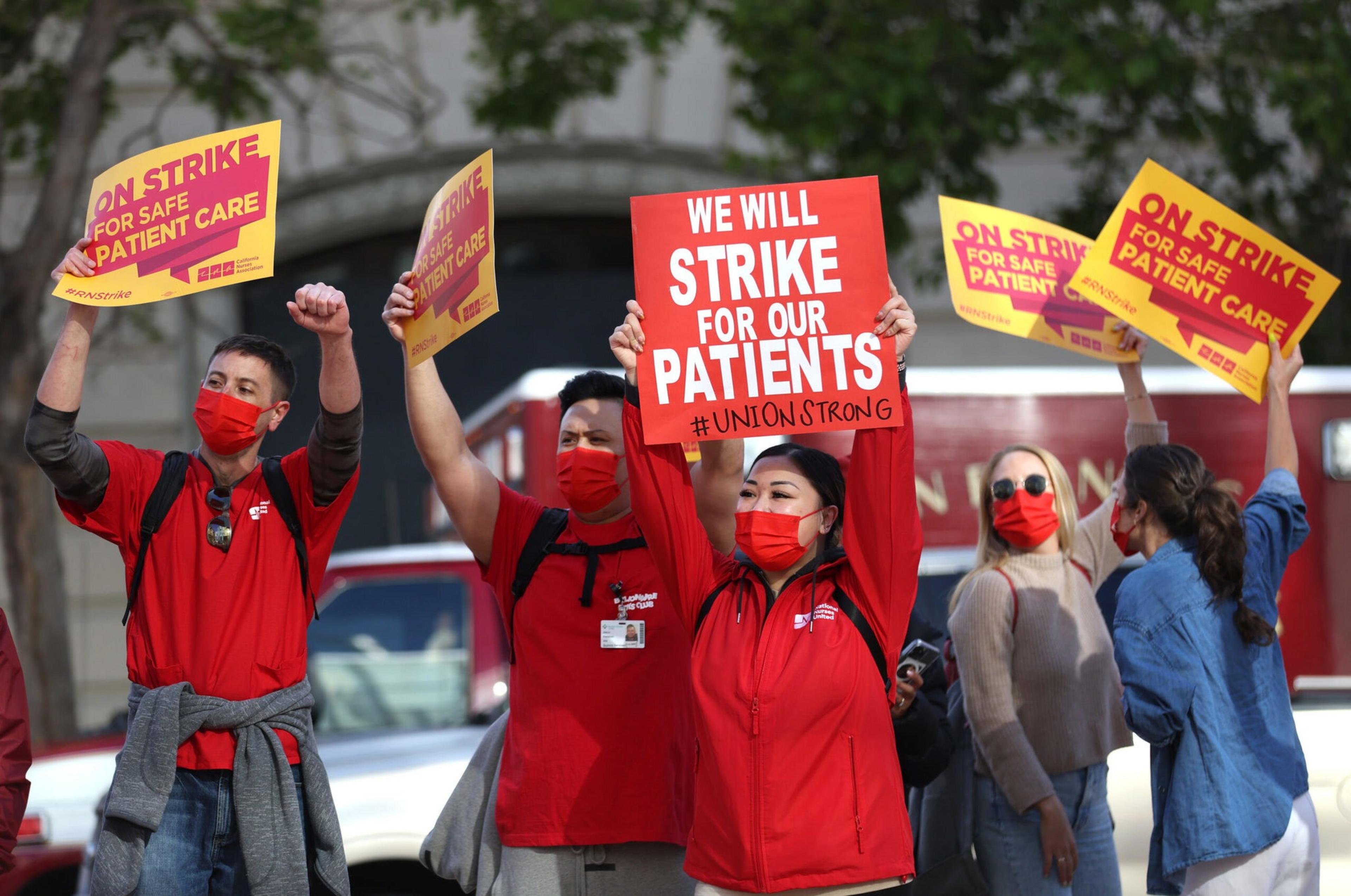Healthcare workers wearing red and holding signs that say "On strike for safe patient care" and "We will strike for our patients" during a protest.