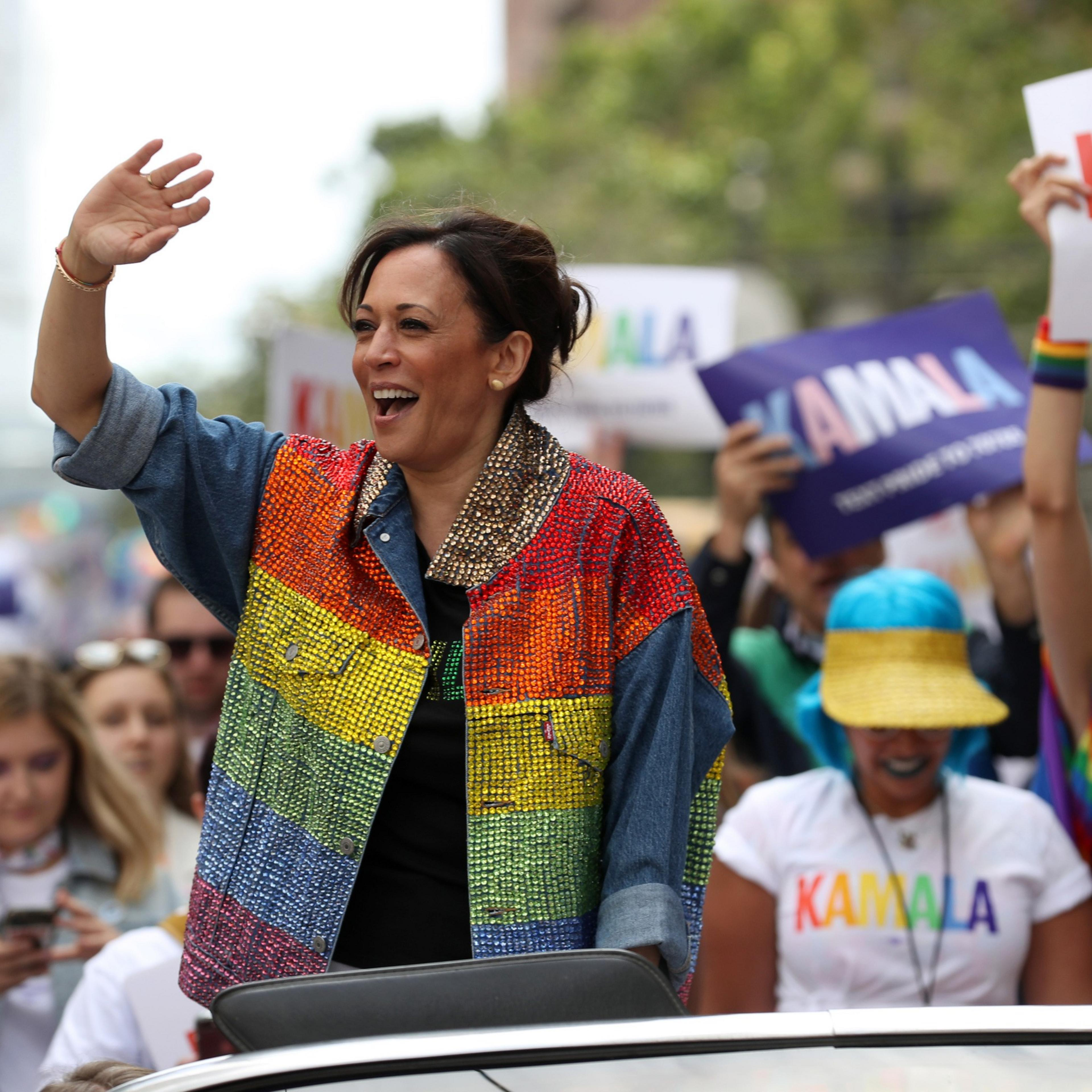 Vice President Kamala Harris waves to crowds wearing a rainbow sequin jacket while standing in a car in a parade.