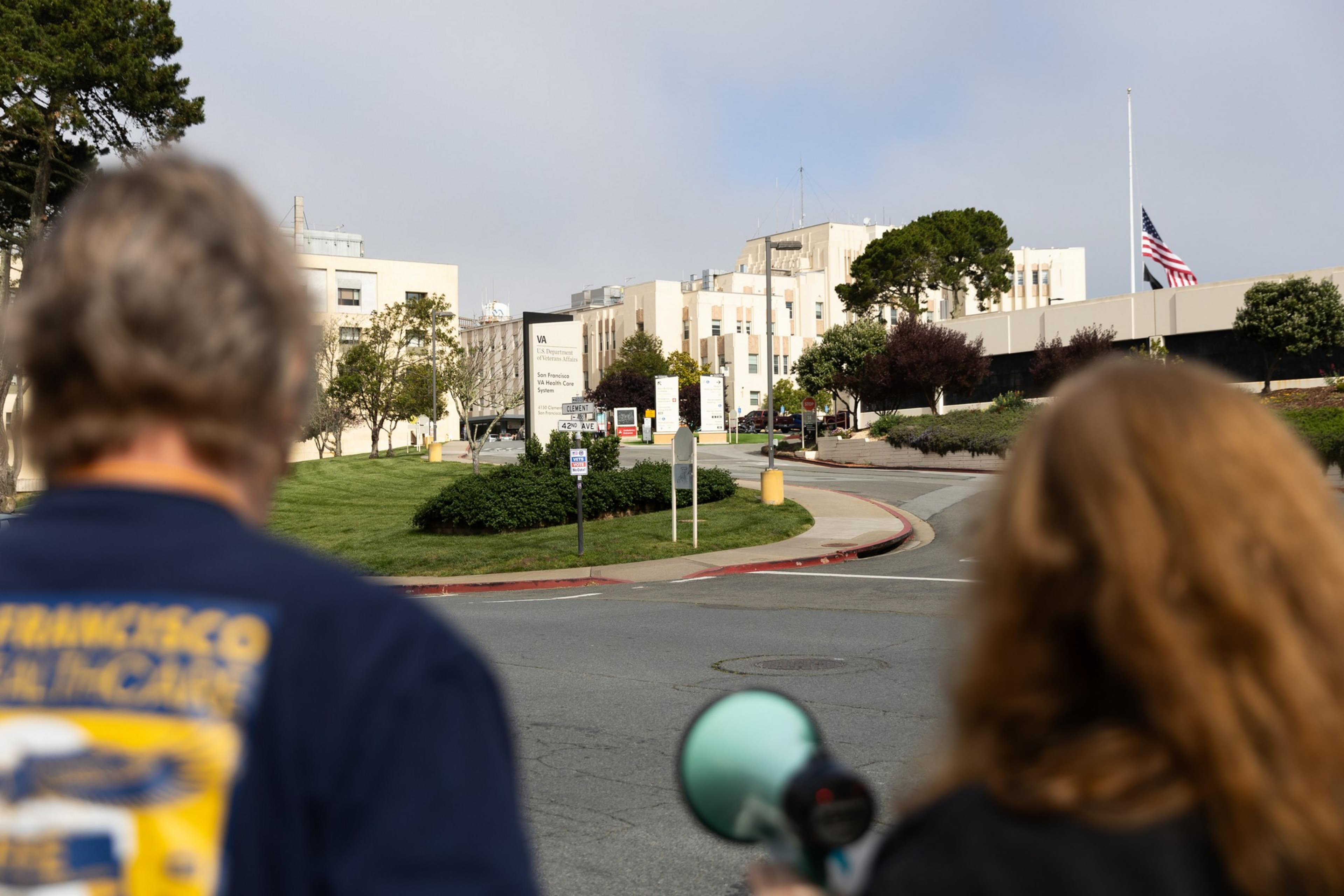 Two people with blurred heads stand on a road, looking toward a hospital entrance. A flag is at half-mast, and signs indicate it's a VA medical center.