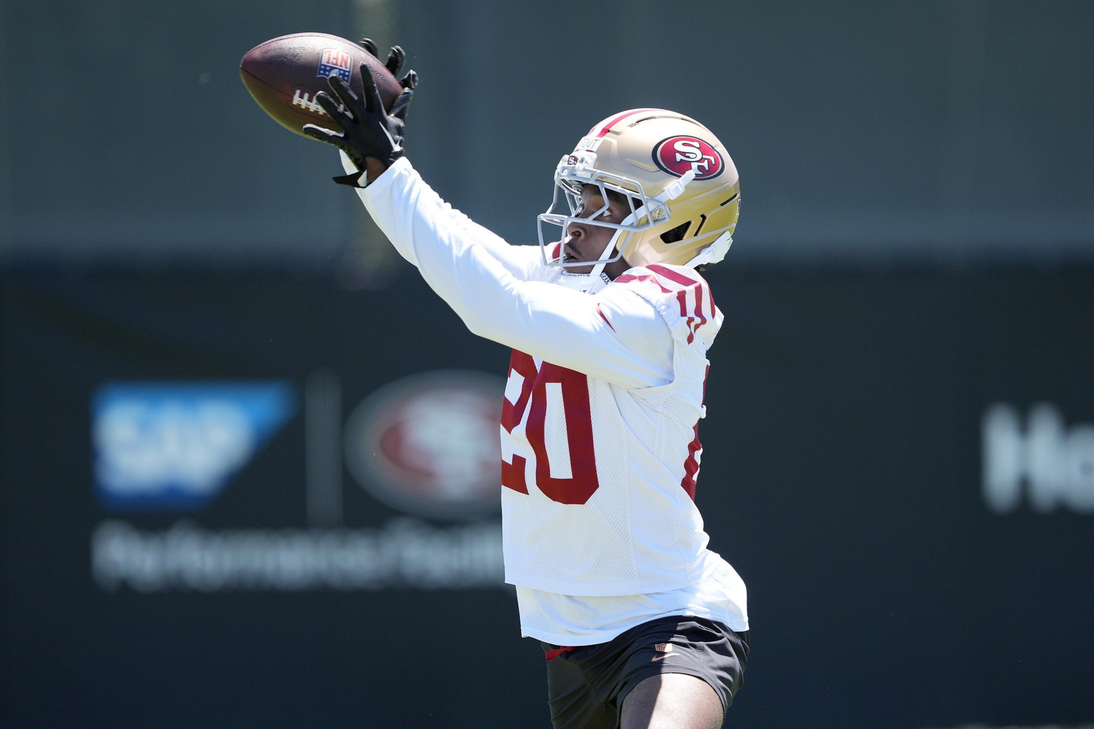 A football player in a red and gold helmet and white jersey is catching a football with both hands during practice, with a blurred background.