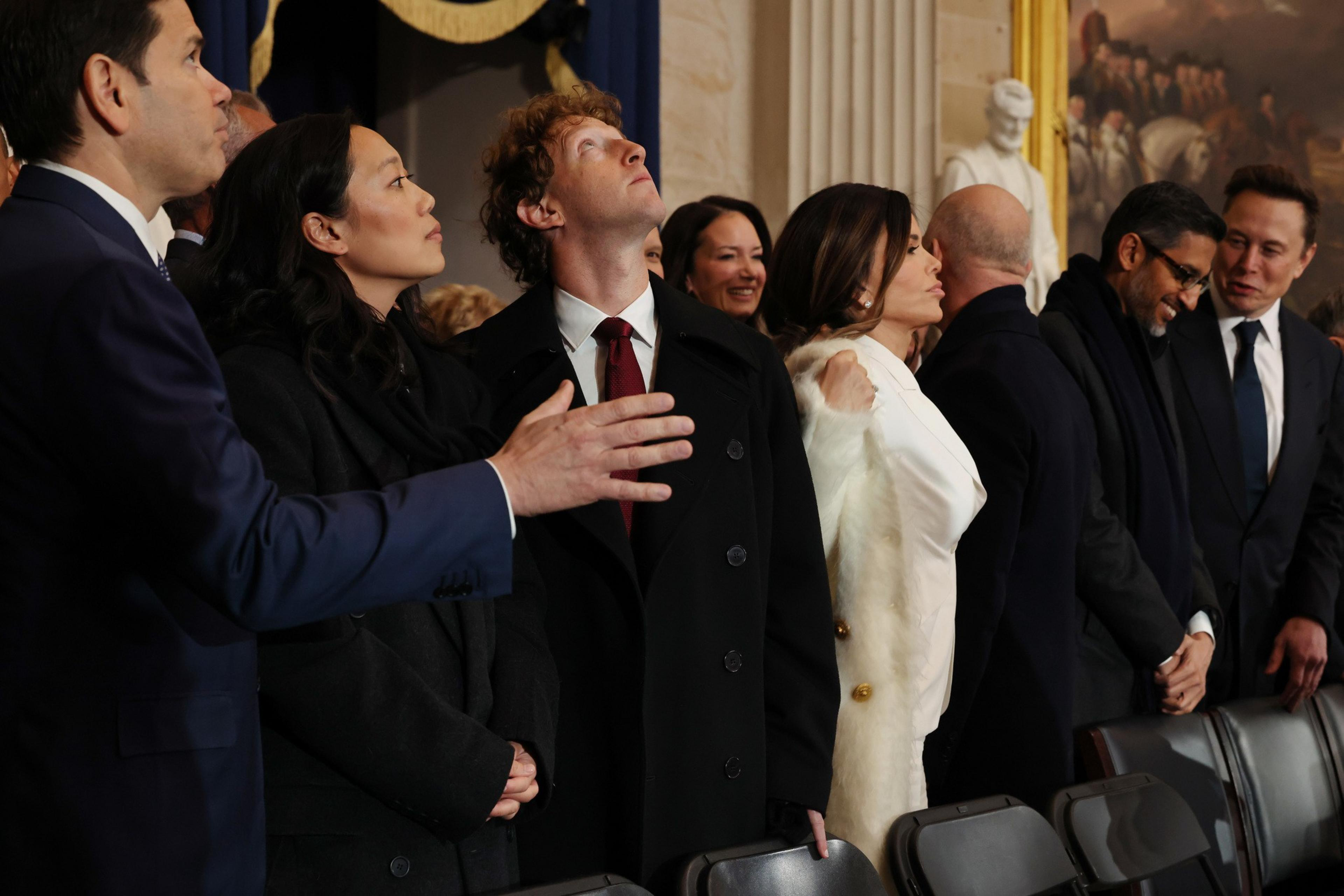 A group of people dressed in formal attire stand together, some looking up. The setting appears to be indoors, with an ornate painting and statue in the background.