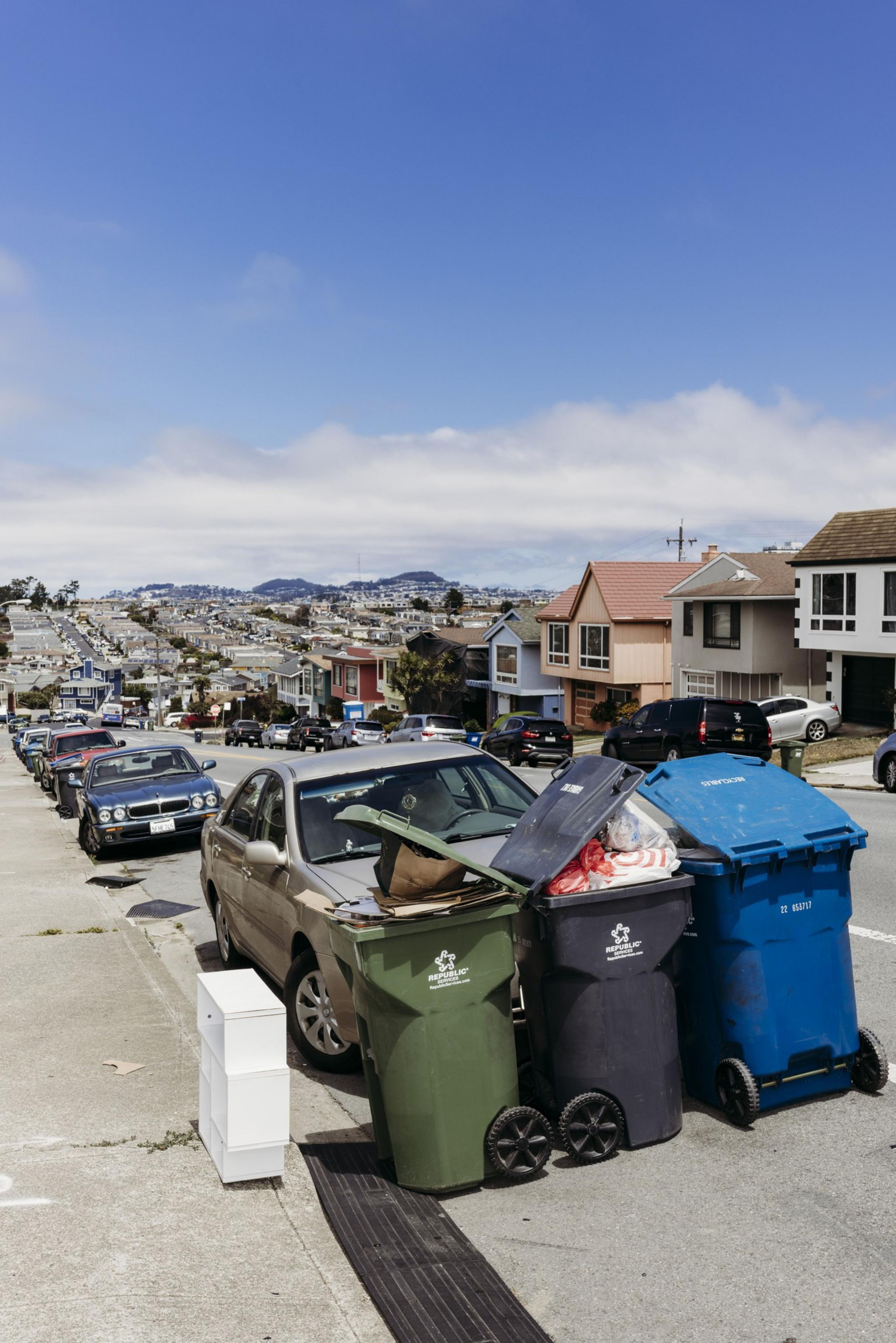 Garbage bins overflowing with trash stand on a residential street lined with parked cars. Houses and hills are visible in the background under a blue sky.