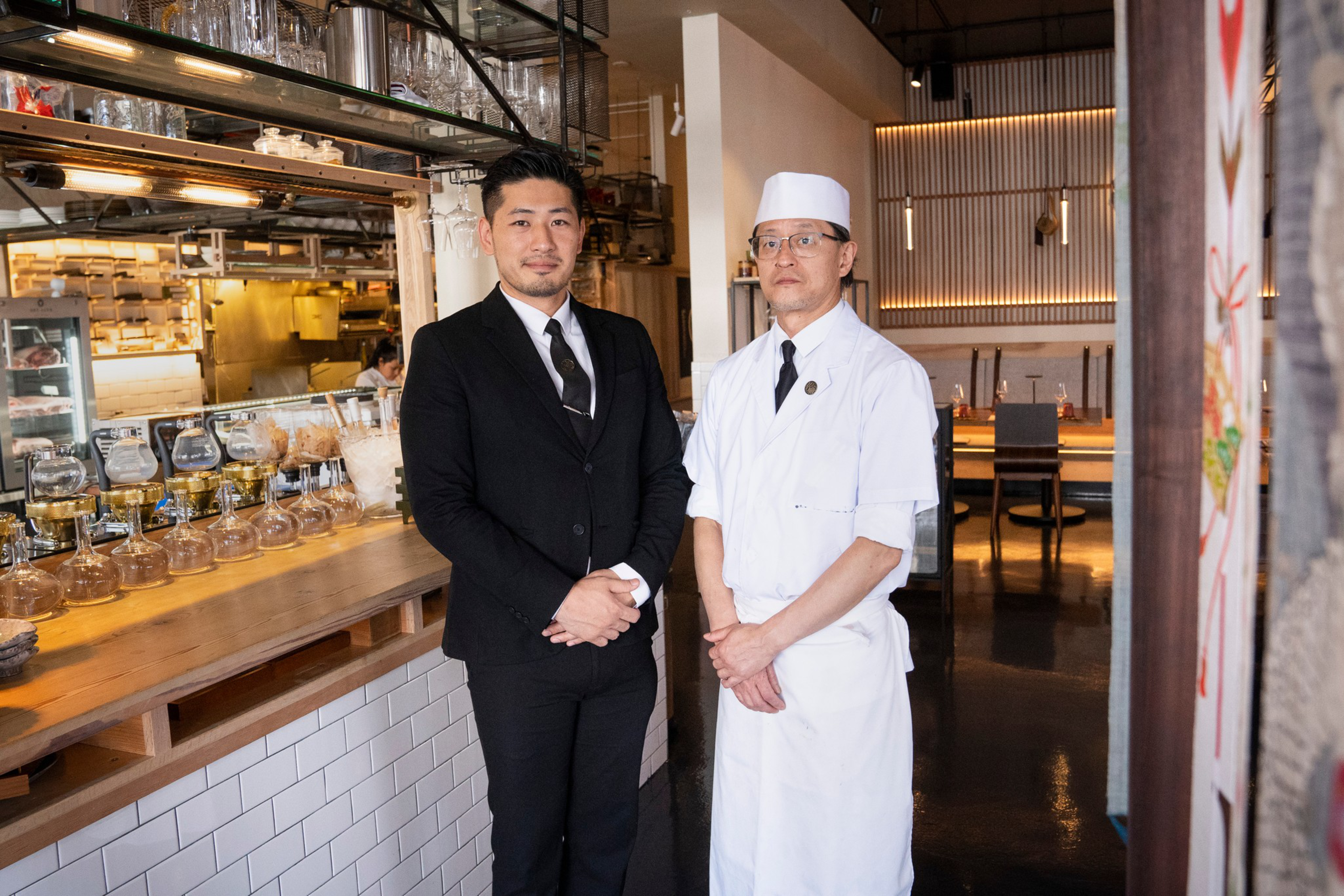 Two men stand in a restaurant, one in a black suit and the other in a white chef's uniform and hat. The bar area is behind them, filled with glassware and equipment.