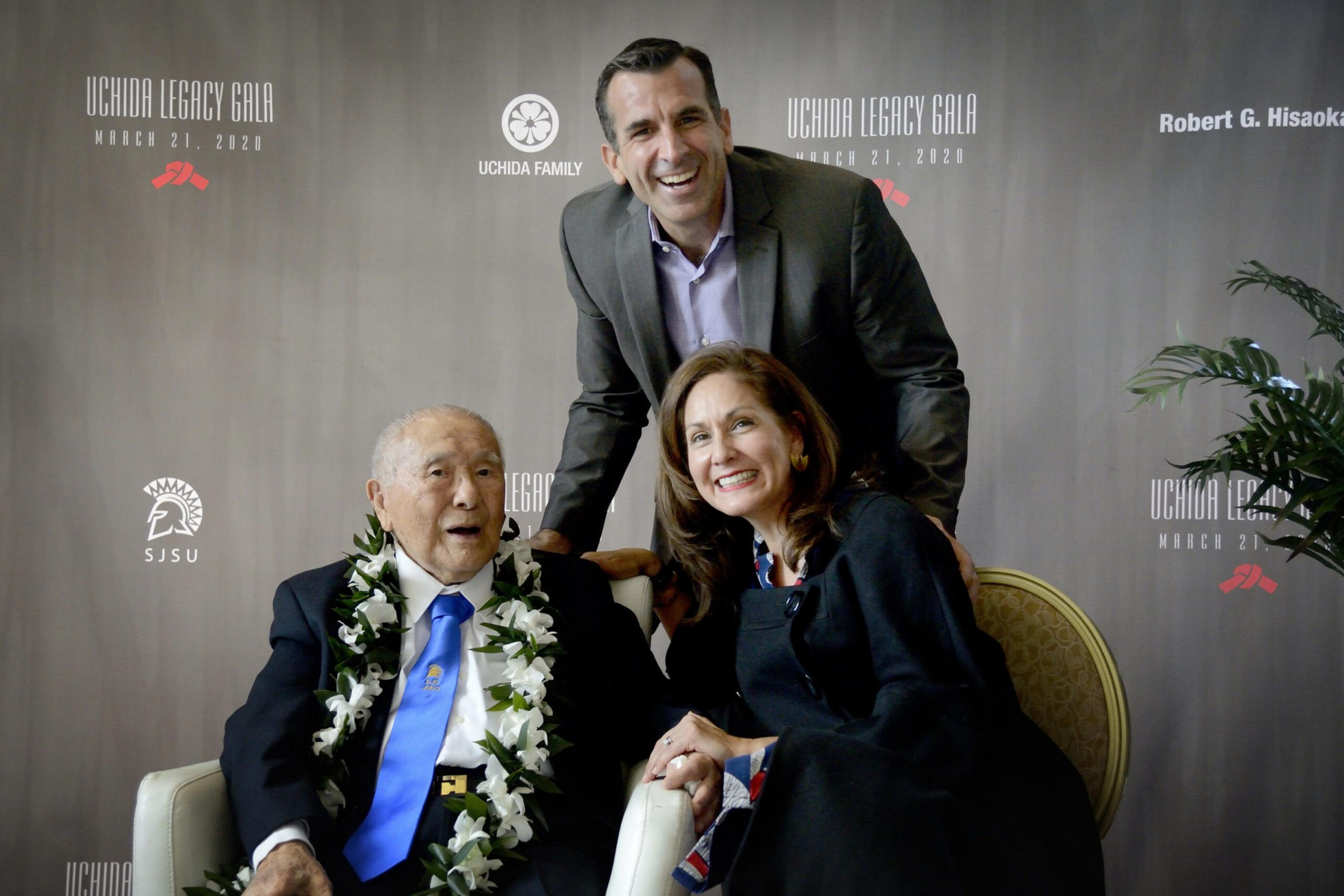 An elderly man with a flower lei, a man in a suit, and a woman in a dark outfit smile together at the Uchida Legacy Gala event backdrop.