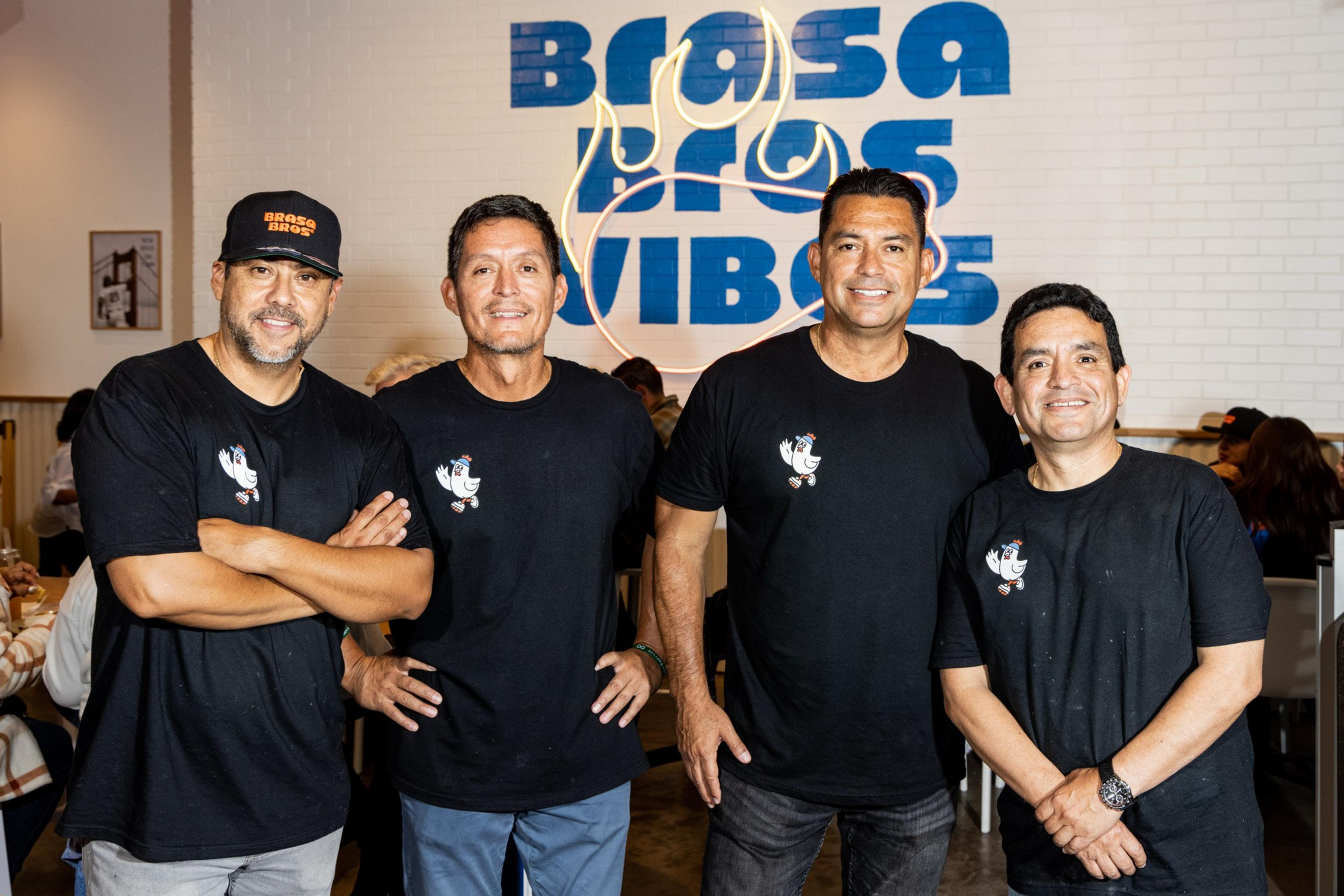 Four men are standing in front of a white brick wall with blue "Brasa Vibes" text and flame logo, all wearing black t-shirts with a small chicken graphic.