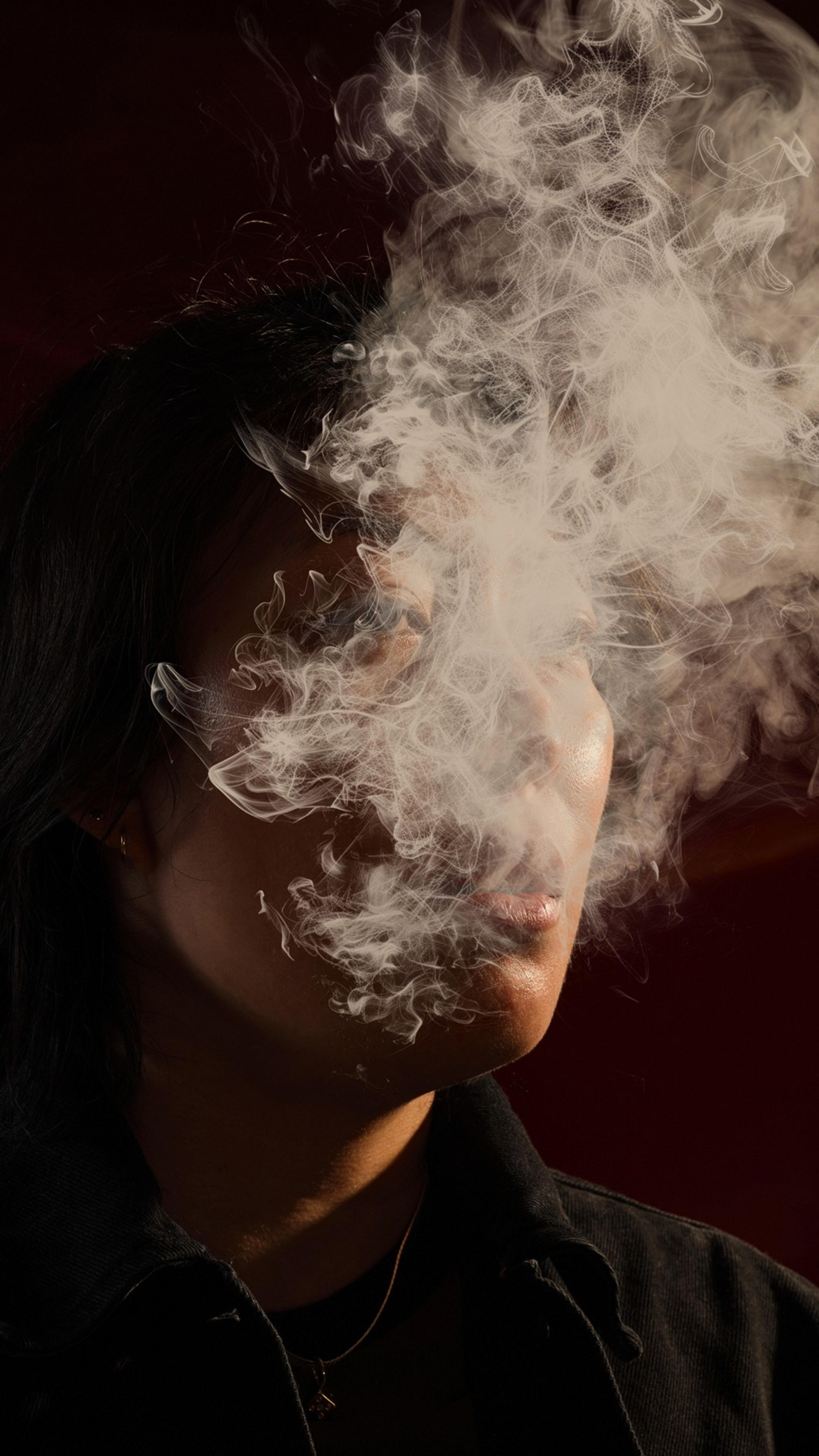 A person with dark hair is partially obscured by a thick cloud of smoke, creating a dramatic and mysterious effect against a dark background.