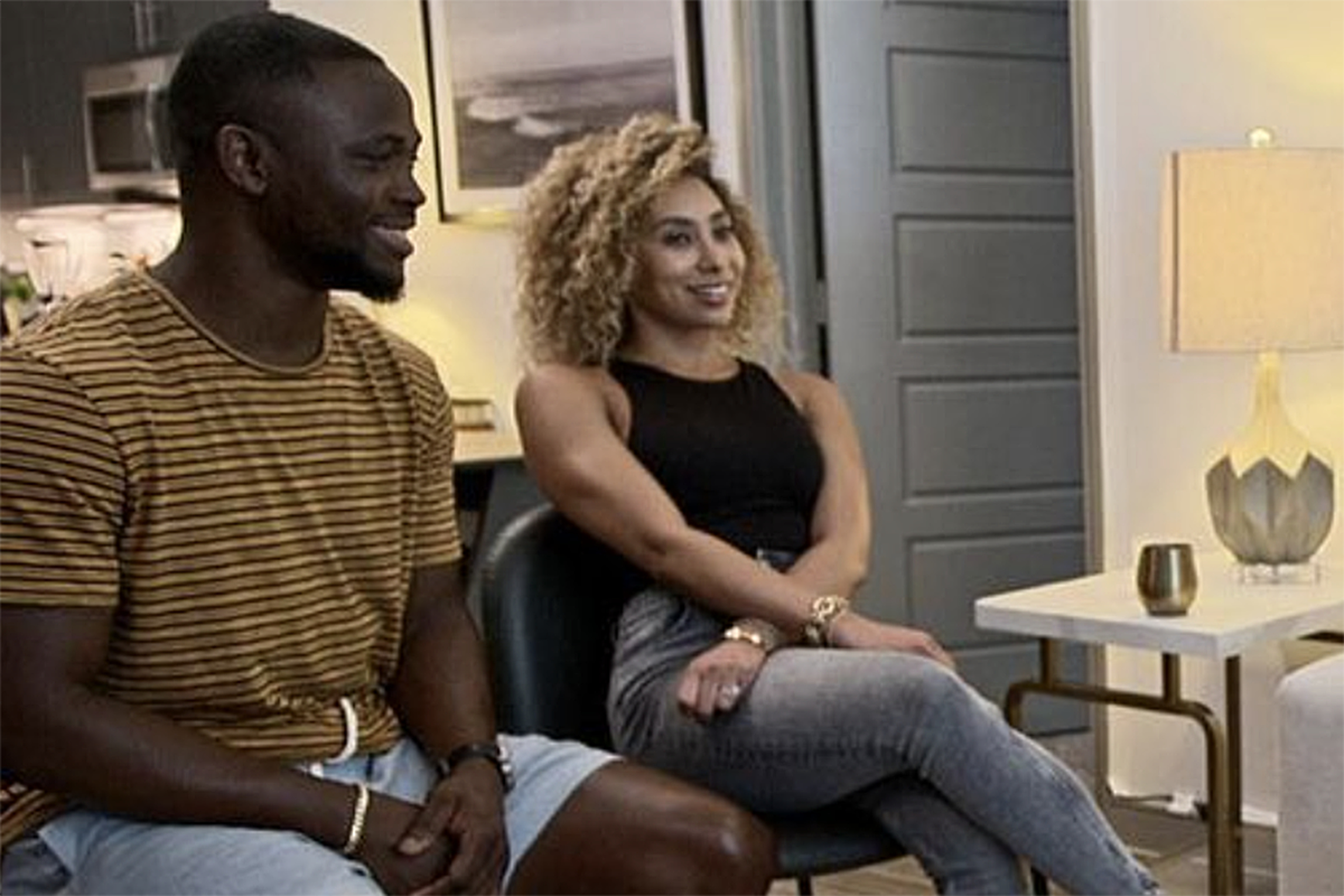 A smiling man in a striped shirt and a woman with curly hair in a black top and jeans sit together in a modern, warmly lit room.