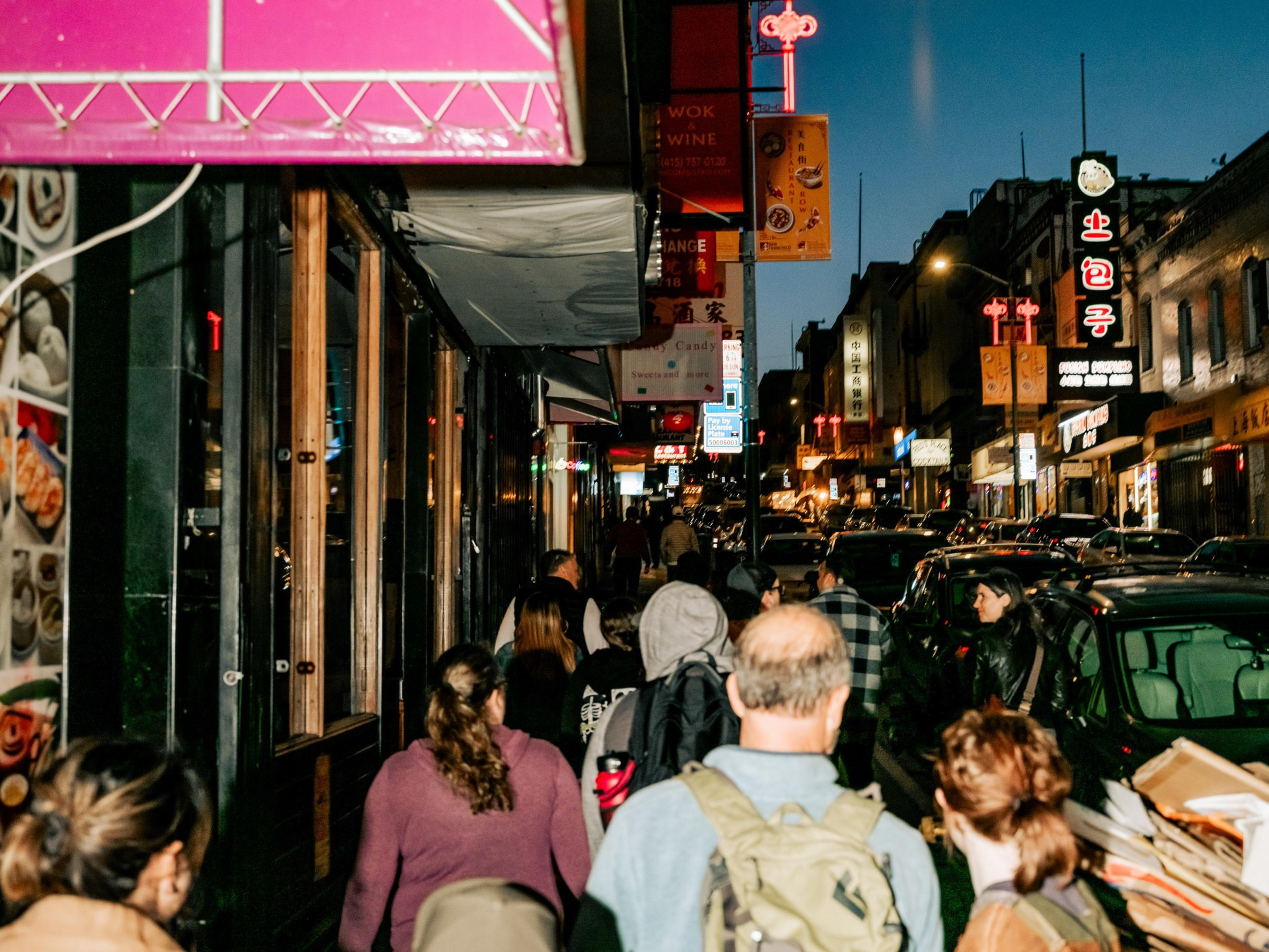 Busy street at night with people walking along the sidewalk. Neon signs light up the scene, and cars are lined up along the street.