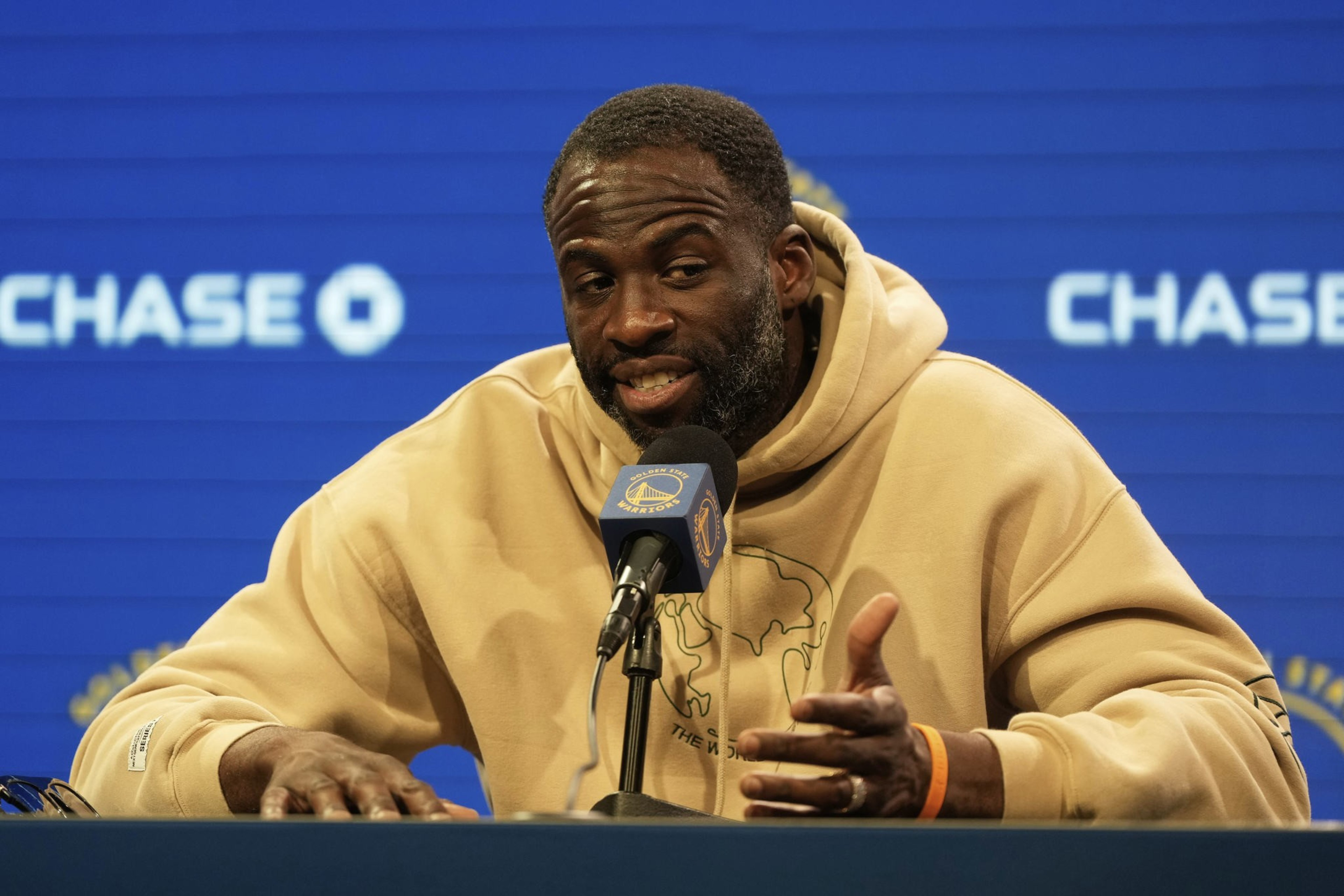 A man in a beige hoodie speaks into a microphone at a press conference. The backdrop is blue with &quot;CHASE&quot; logos.