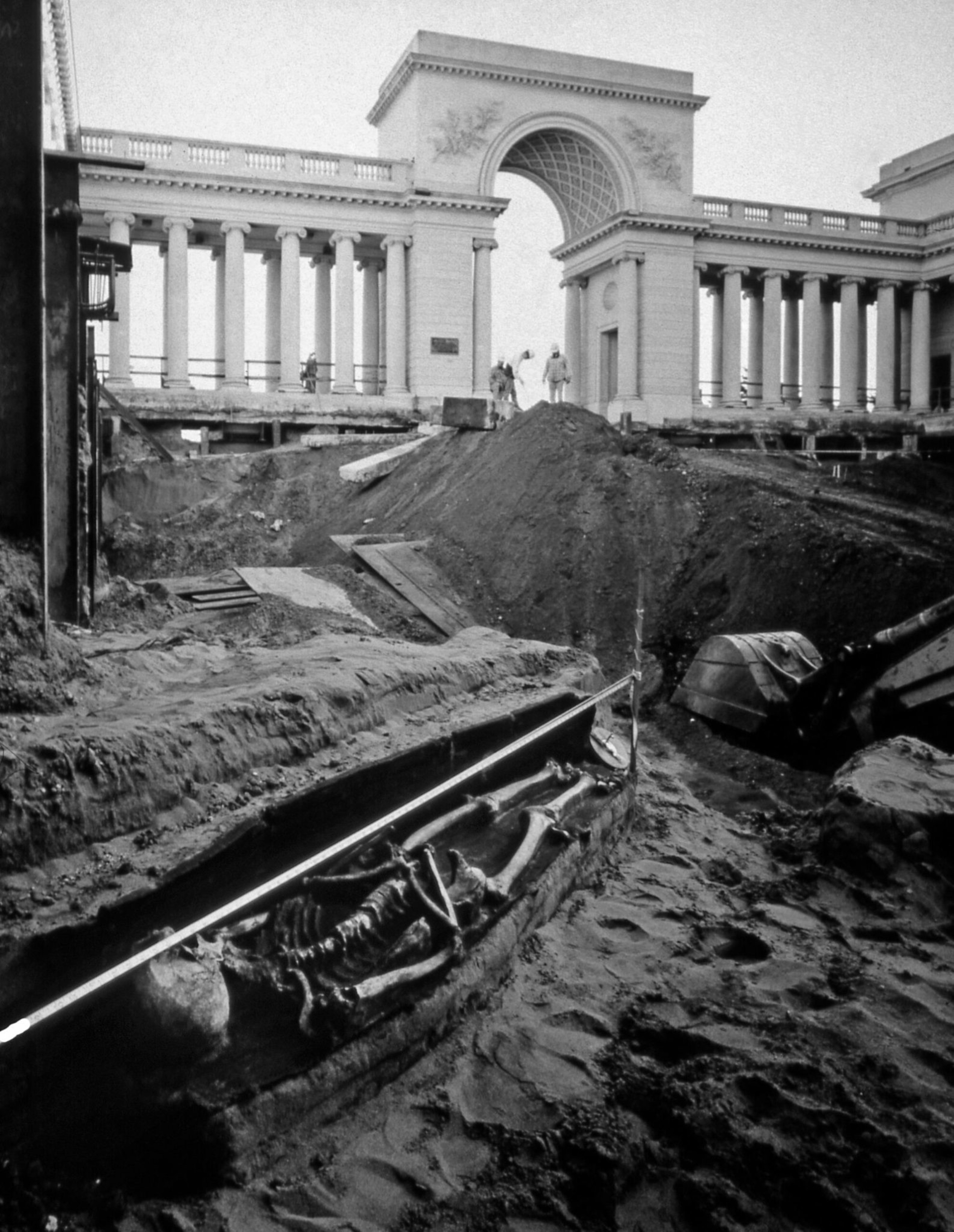 A skeleton lies partially uncovered in a trench with construction workers and a grand columned archway in the background.