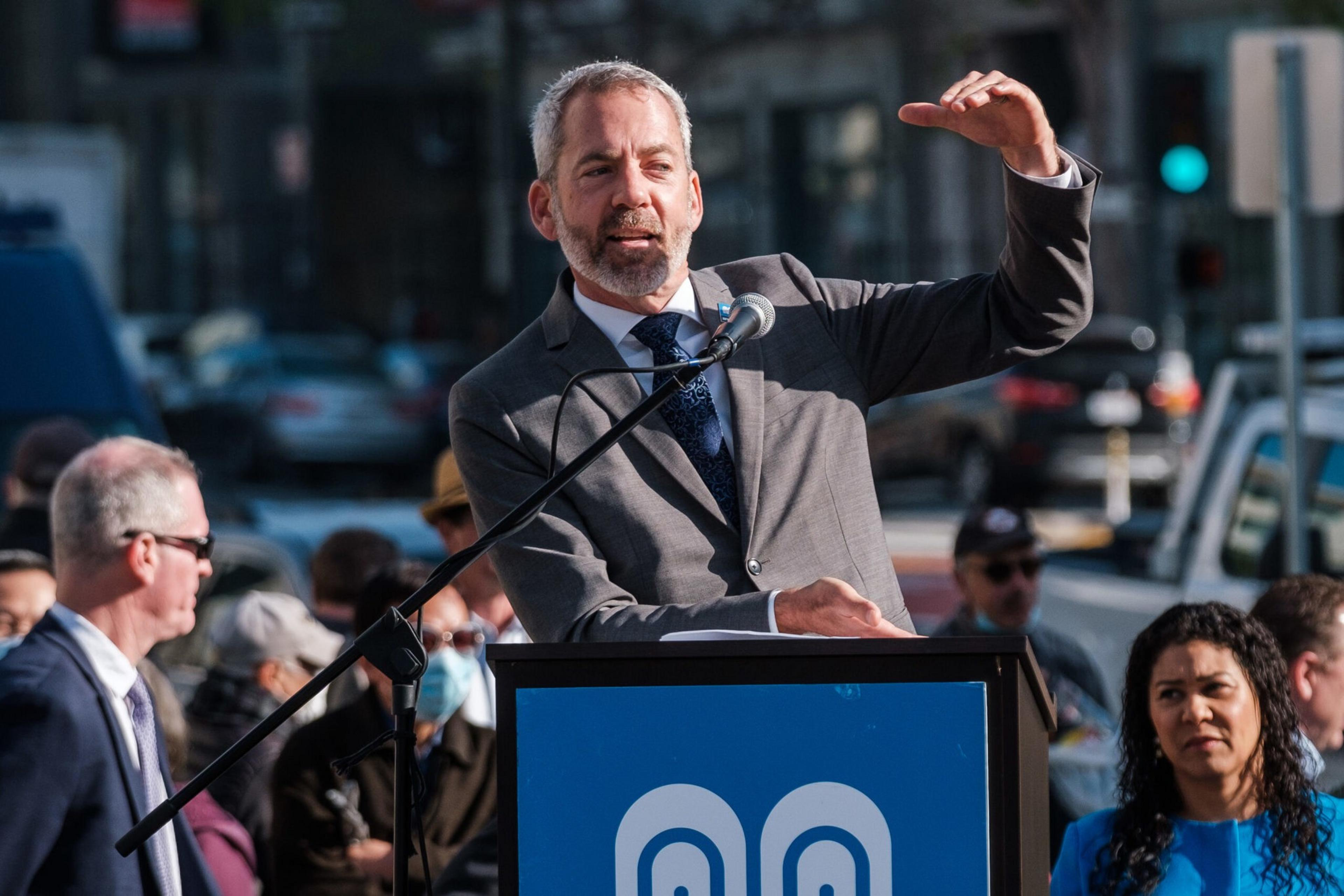 A man in a suit speaks at a podium with a microphone, gesturing with his right hand, while a crowd watches in the background.