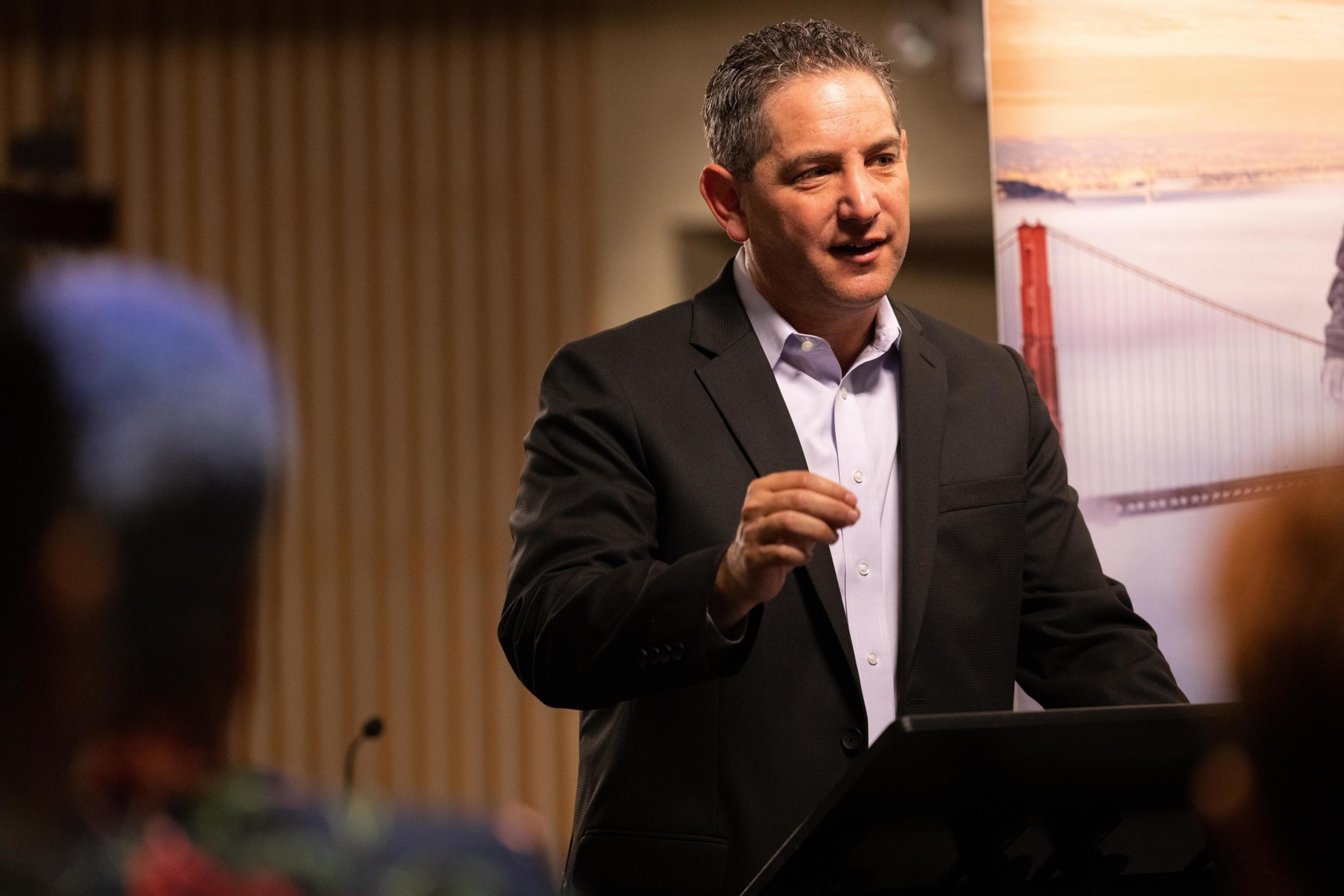 A man in a suit is speaking at a podium, gesturing with one hand. Behind him is a poster with part of a bridge visible. The setting appears to be indoors.
