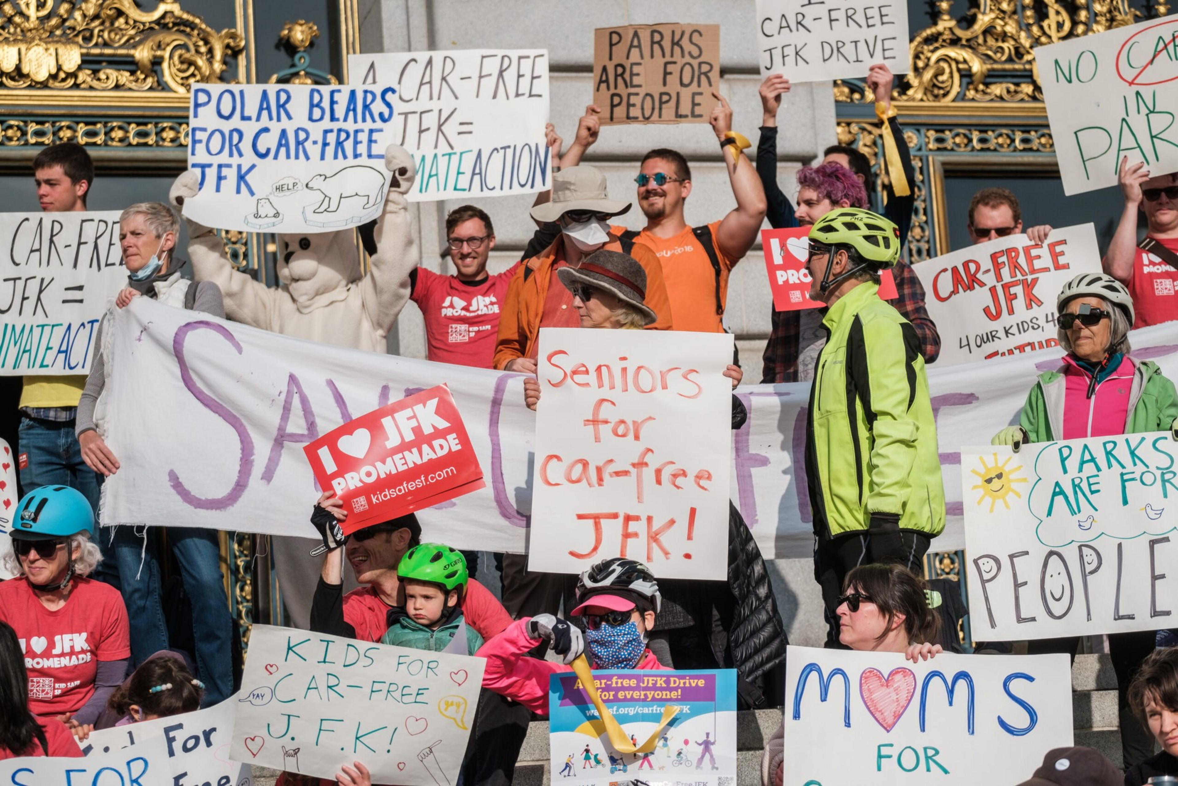 People hold signs advocating for a car-free JFK Drive, with messages about parks, seniors, kids, and polar bears supporting the cause.