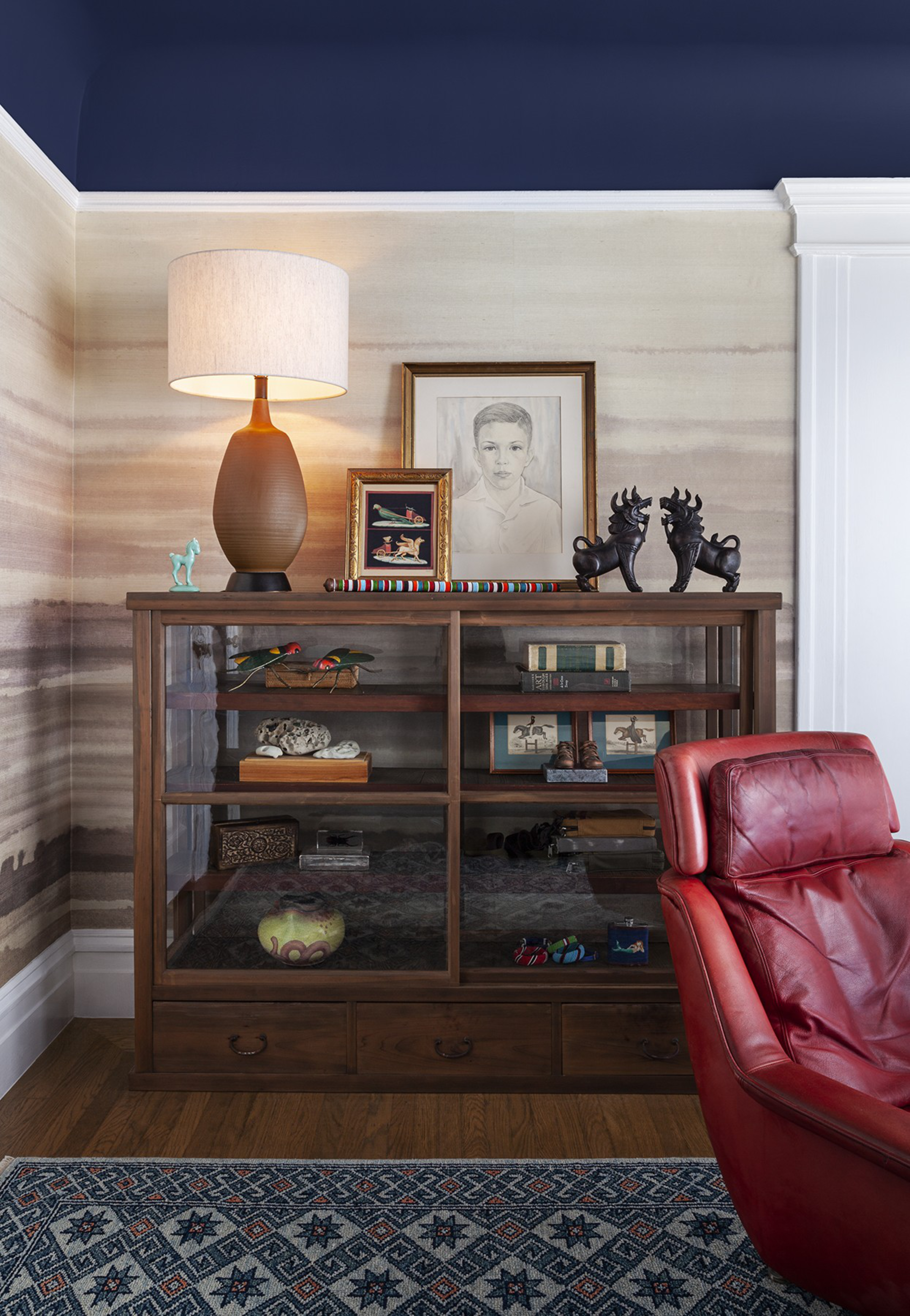 A wooden cabinet with art, books, and decor is topped with a lamp, framed drawings, and statues. In front is a red leather chair and a blue patterned rug on the floor.