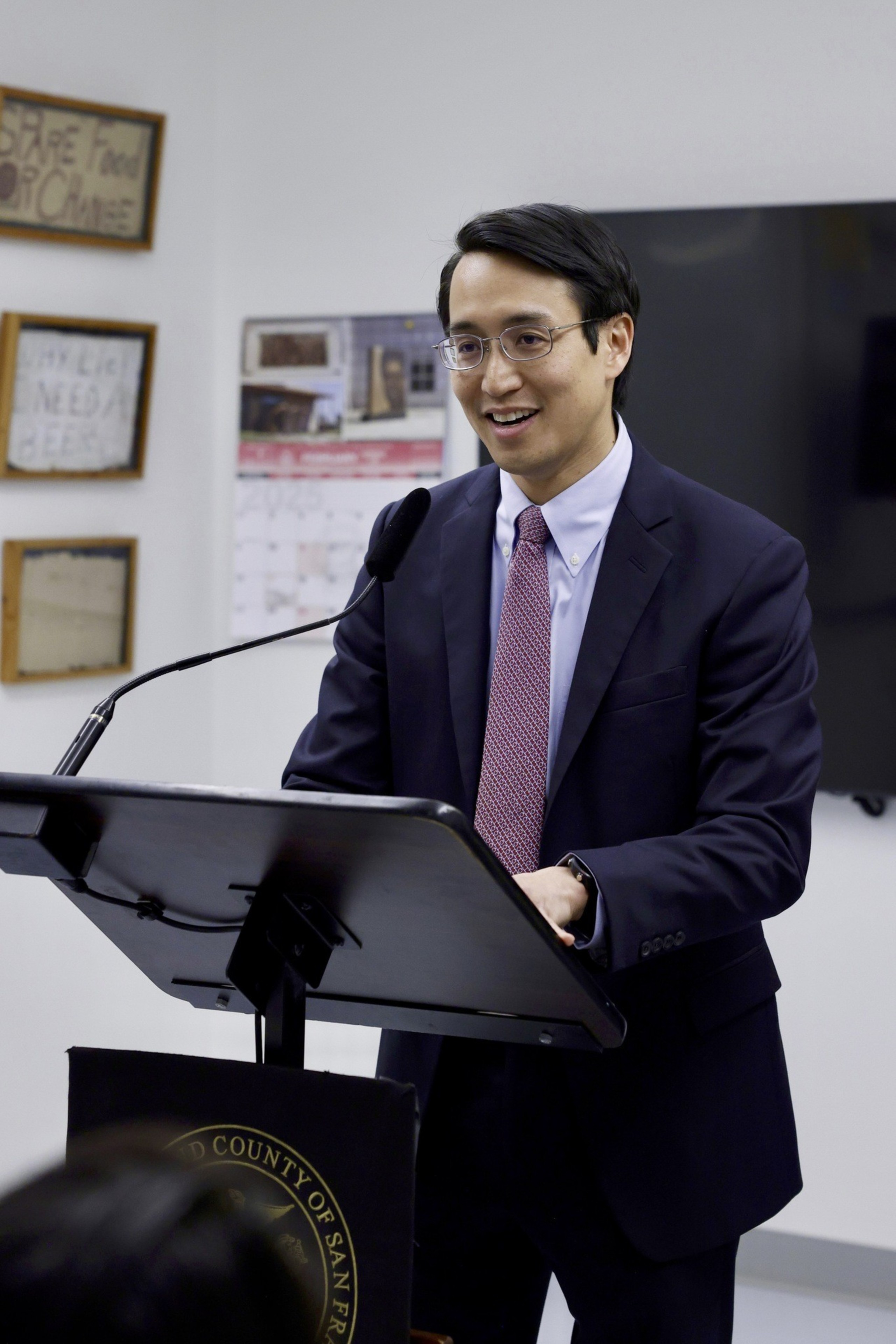 A man in a suit and tie is speaking at a podium with a microphone. There's a calendar and framed signs on the wall behind him.