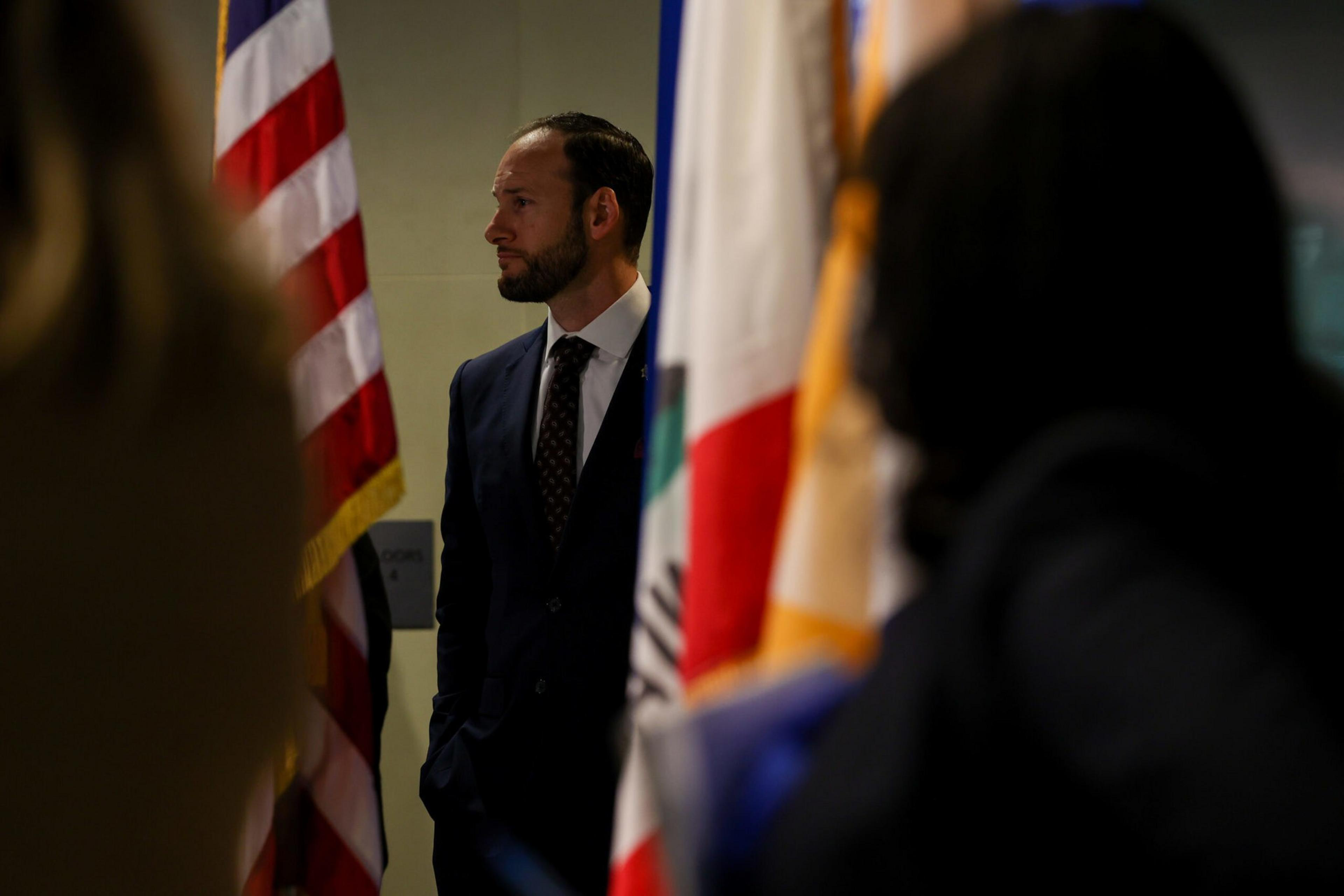 A man in a dark suit stands with hands in pockets, surrounded by blurred flags, facing left with an American flag partially visible behind him.
