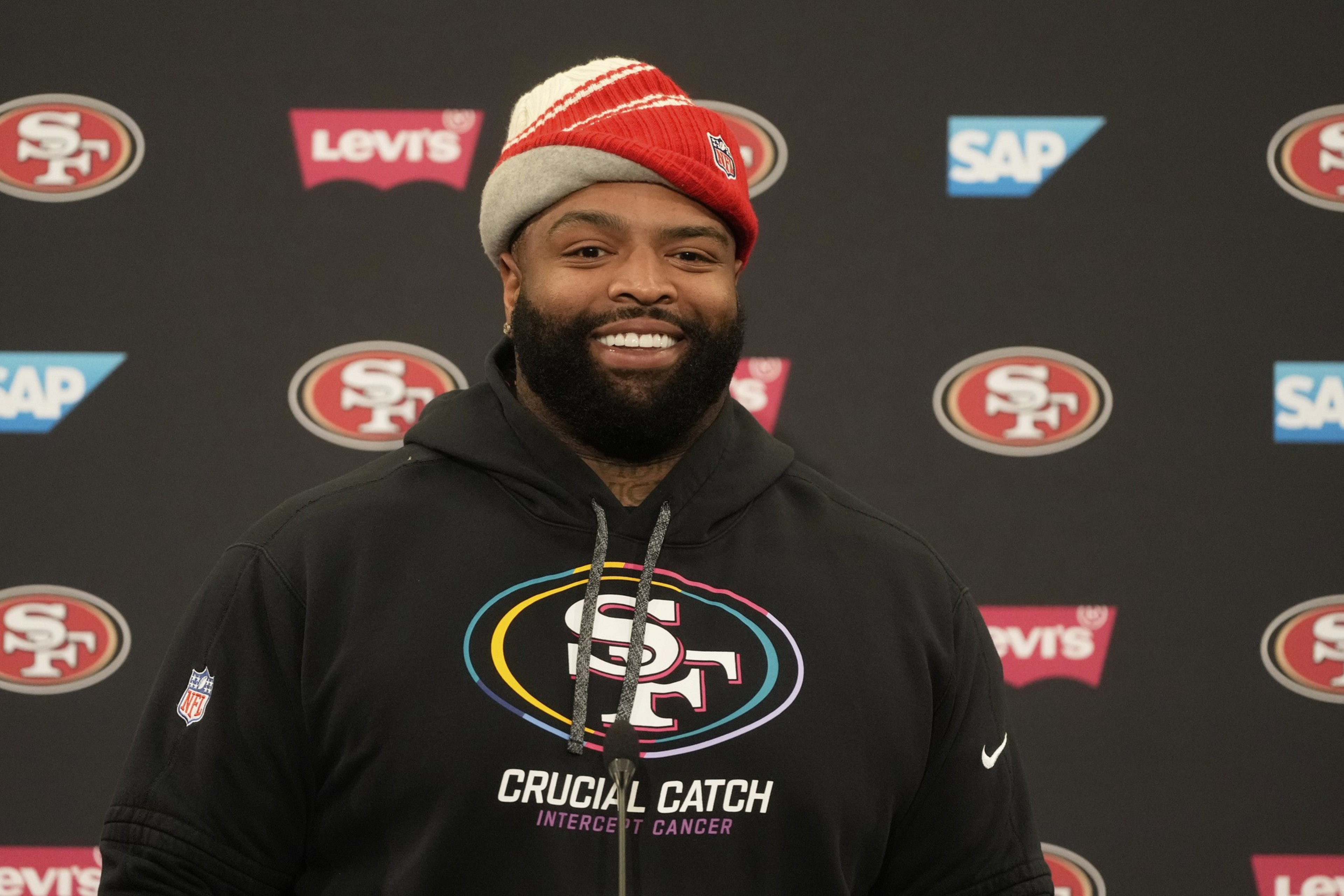 A smiling man in a black hoodie and red beanie stands against a backdrop with 49ers and brand logos. His hoodie reads "Crucial Catch, Intercept Cancer."