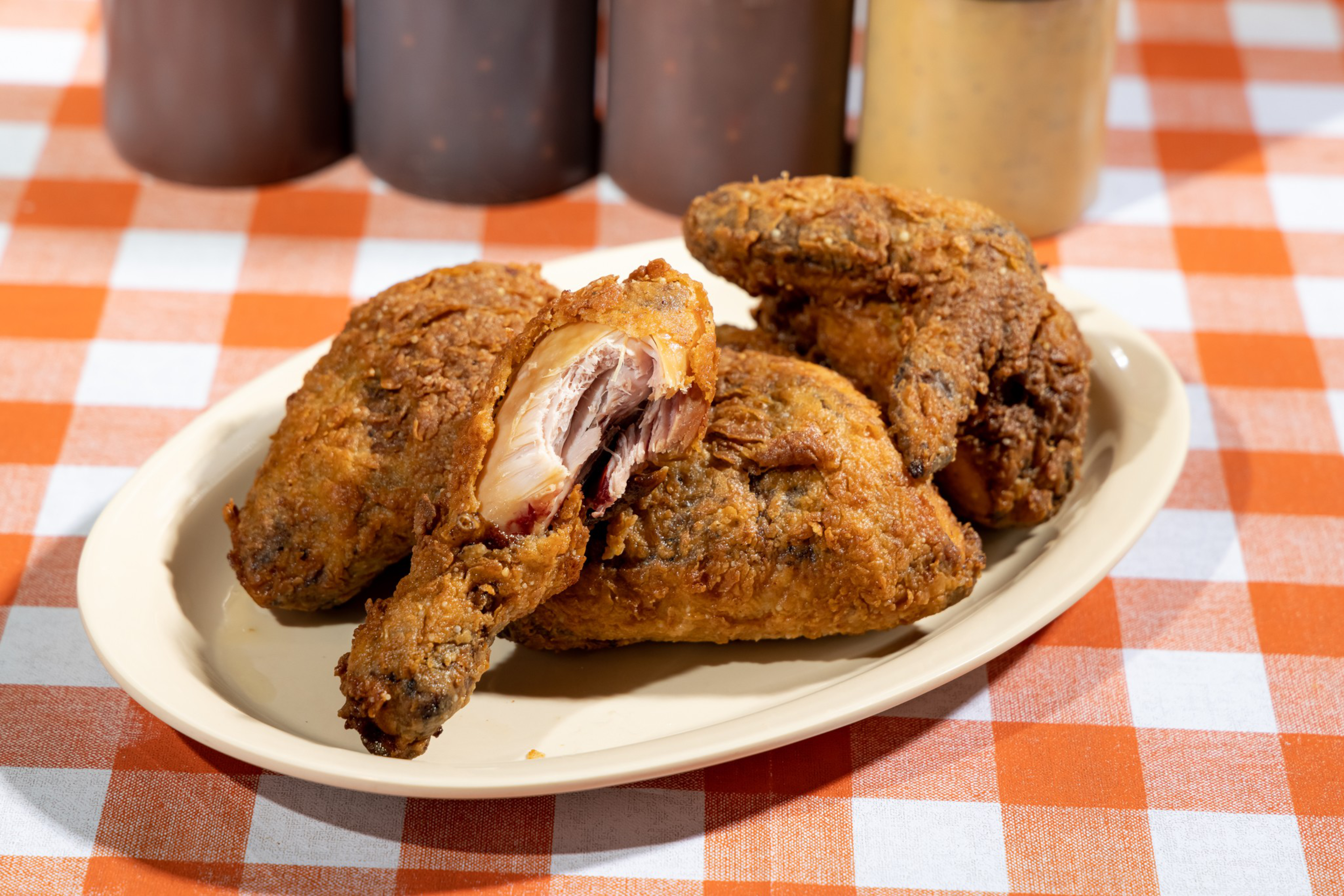 Three pieces of crispy fried chicken, one partially eaten, are served on a white oval plate on an orange and white checkered tablecloth.