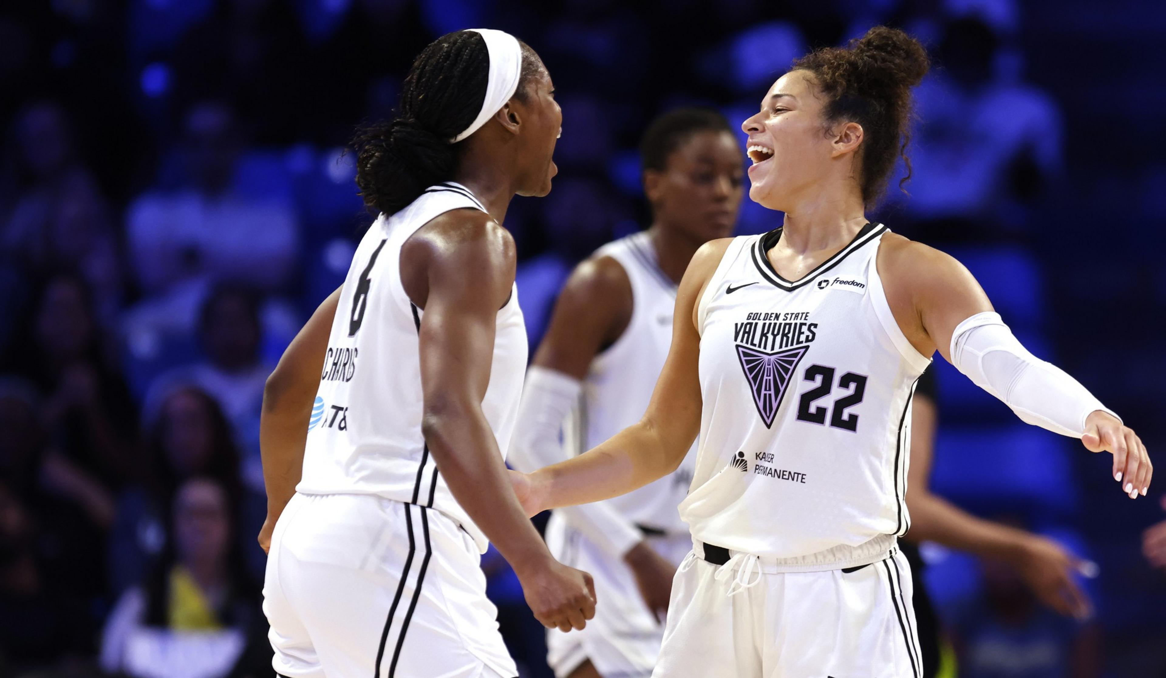 Two female basketball players in white Golden State Valkyries uniforms are celebrating, smiling and reaching out to each other energetically on the court.