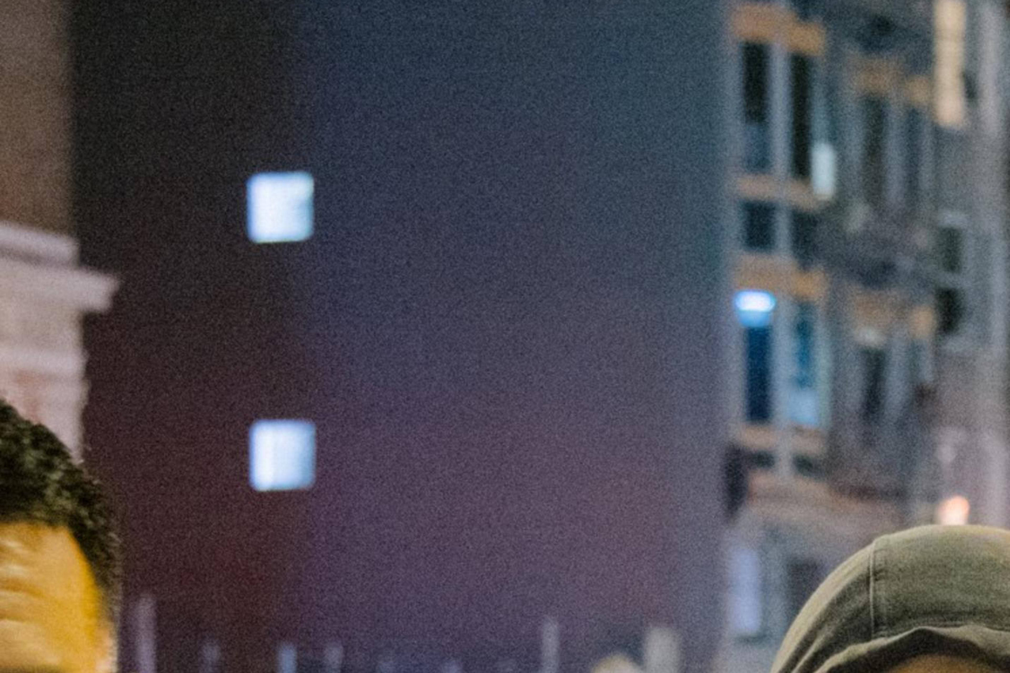 A woman in a hooded jacket stands on a bustling city street at night, with illuminated buildings and tents in the background.
