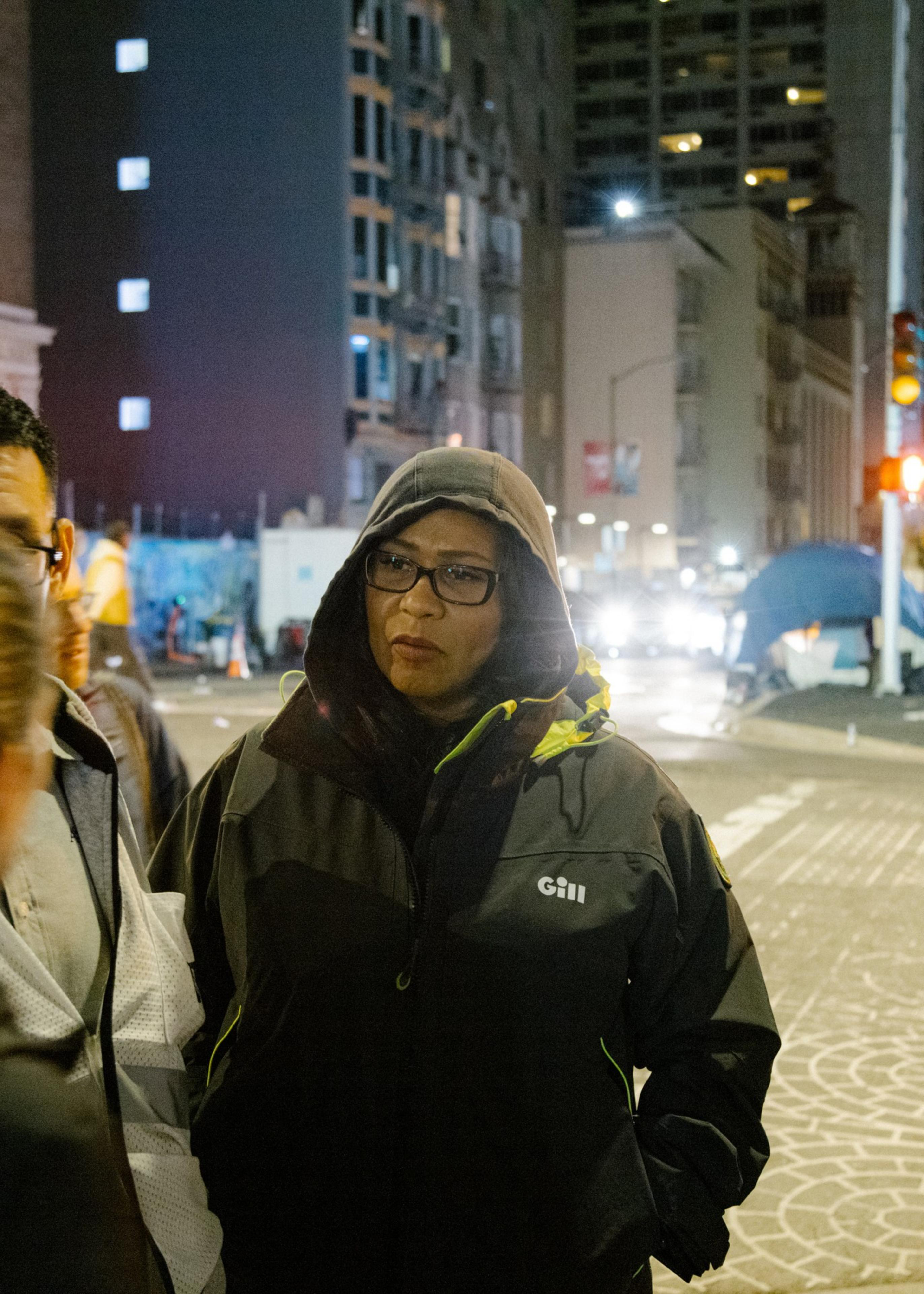 A woman in a hooded jacket stands on a bustling city street at night, with illuminated buildings and tents in the background.