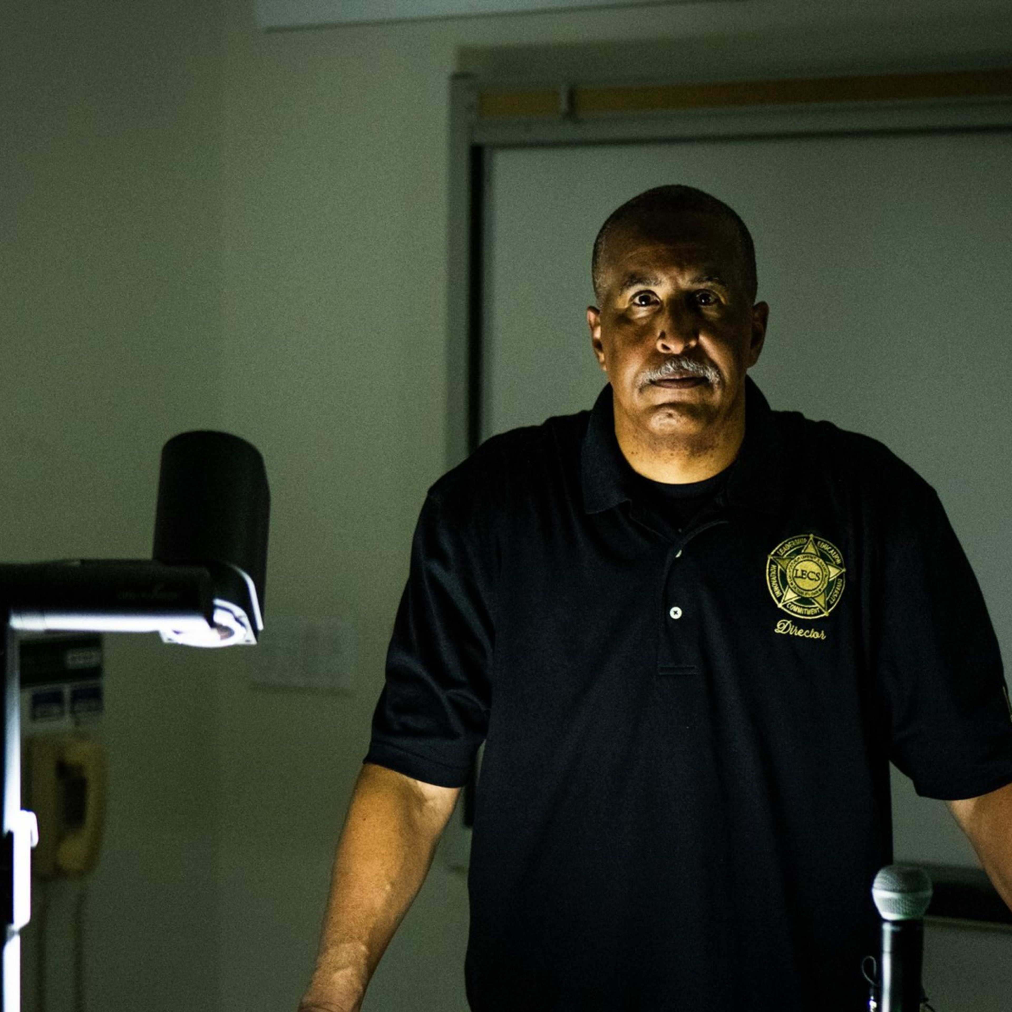 A man in a dark polo shirt stands at a lectern with a computer and a document camera, lit dramatically from above, against a dimly lit classroom backdrop.